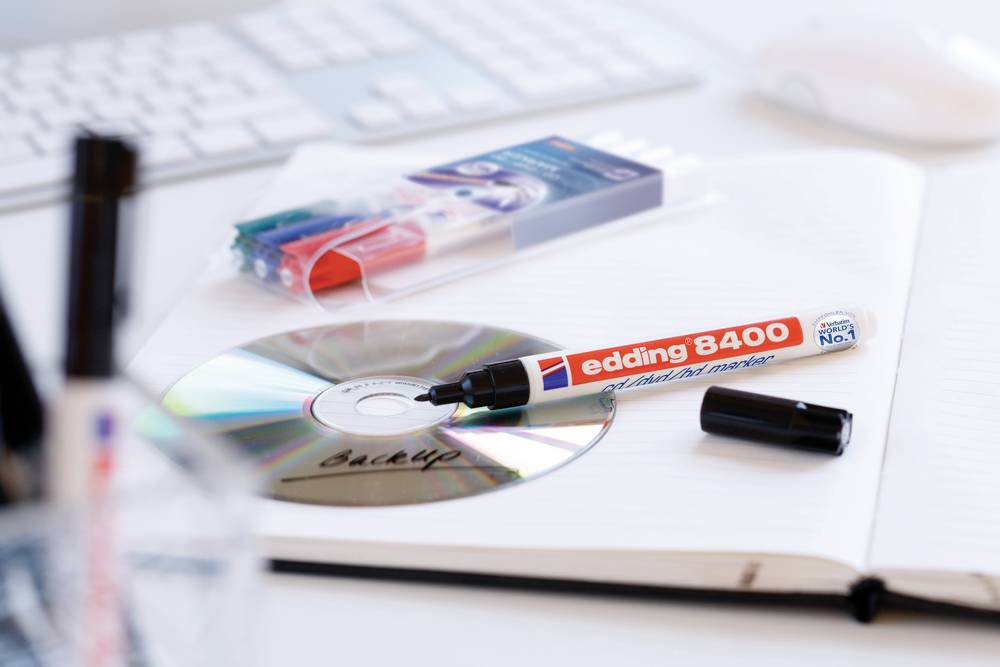 Ein roter Marker 'edding 8400' liegt auf einem offenen Notizbuch und einer beschrifteten CD. Im Hintergrund eine Tastatur und weitere Stifte.