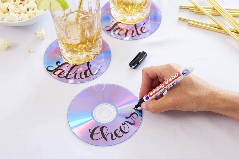 Eine Hand beschriftet glänzende CDs mit einem Marker, die als Untersetzer dienen. Zwei Gläser mit Getränken stehen auf bereits beschrifteten CDs.