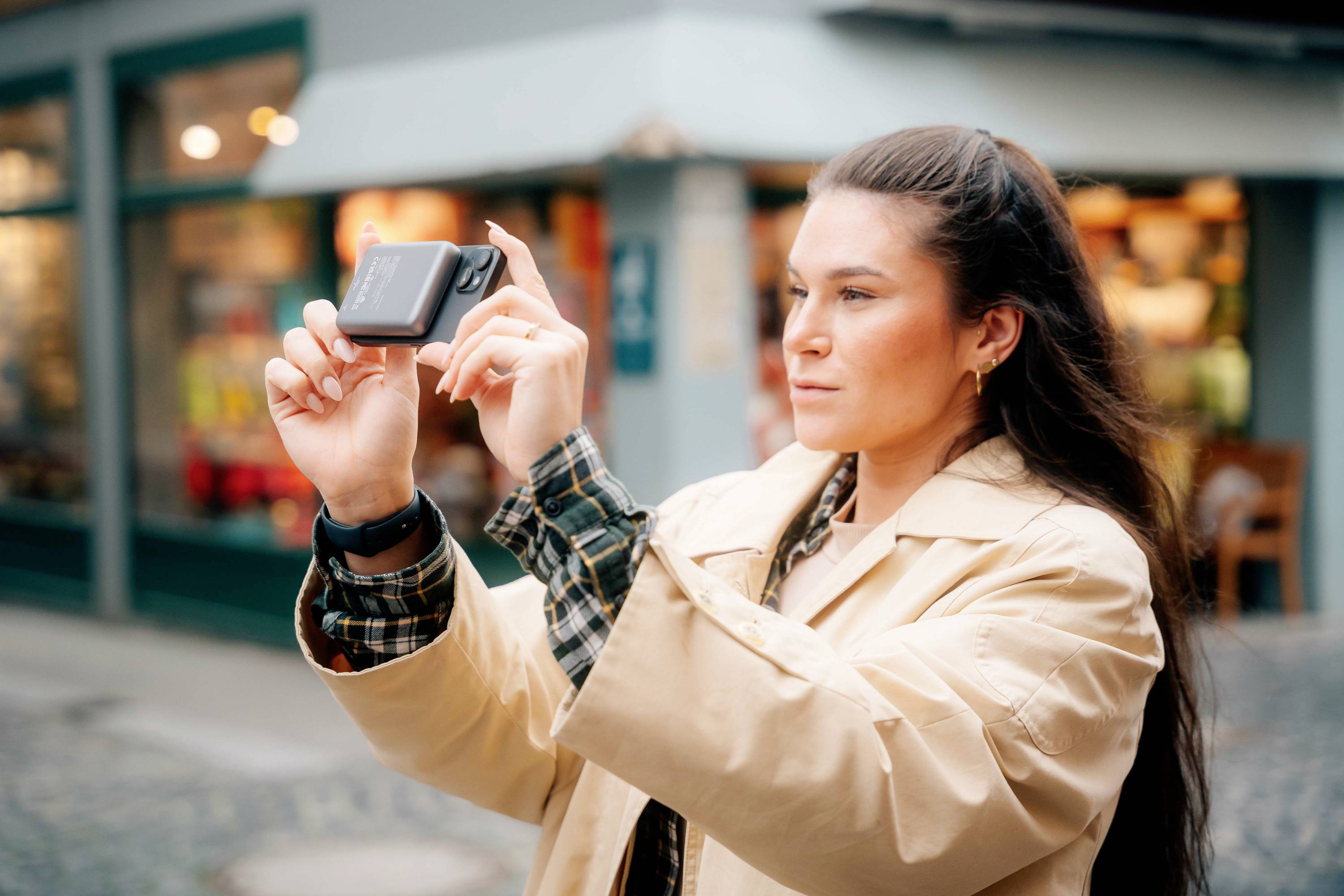 Eine Person hält eine Kamera hoch und macht ein Foto in einer urbanen Umgebung. Im Hintergrund sind Geschäfte zu sehen.