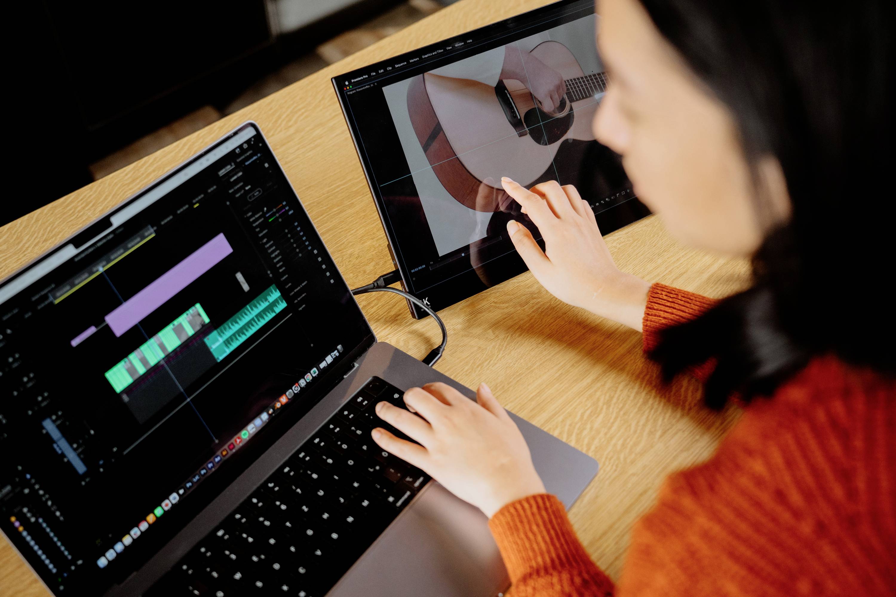 Eine Person arbeitet an einem Laptop und einem Tablet. Auf dem Laptop ist ein Video-Editor mit Tonspuren zu sehen, während das Tablet ein Bild einer Gitarre zeigt.