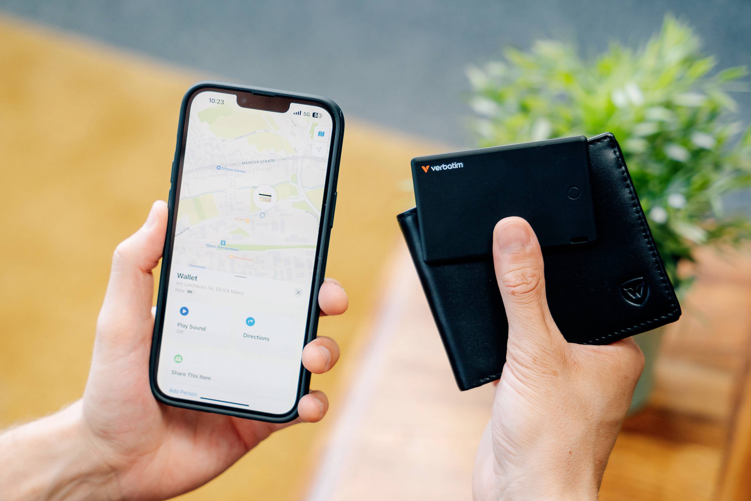 Eine Person hält ein Smartphone mit einer Karten-App in einer Hand und eine schwarze Geldbörse in der anderen, im Hintergrund eine Pflanze.