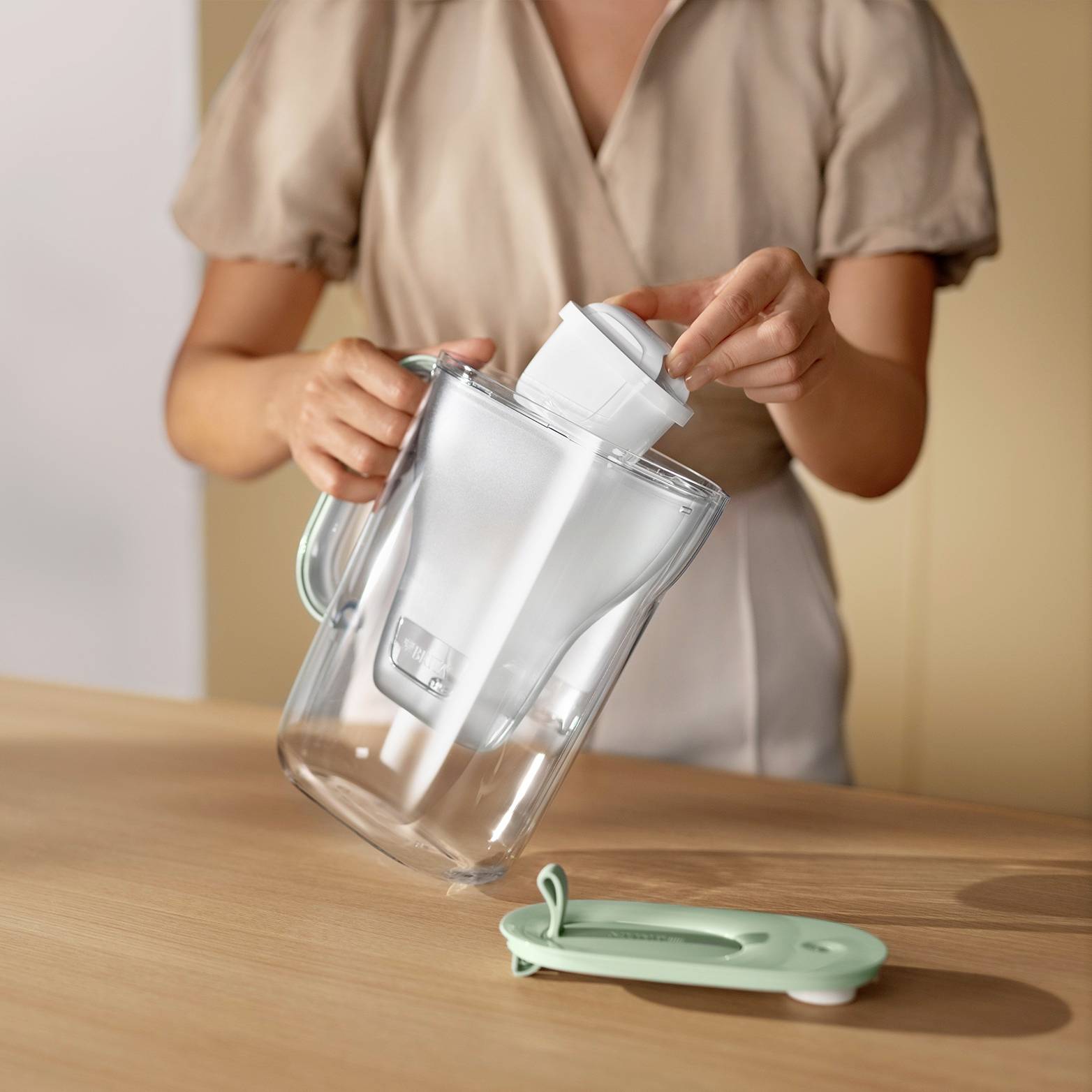 Eine Person gießt Wasser aus einem transparenten Krug mit Filter, der auf einem Holztisch steht. Sie trägt ein beige geknöpftes Hemd.