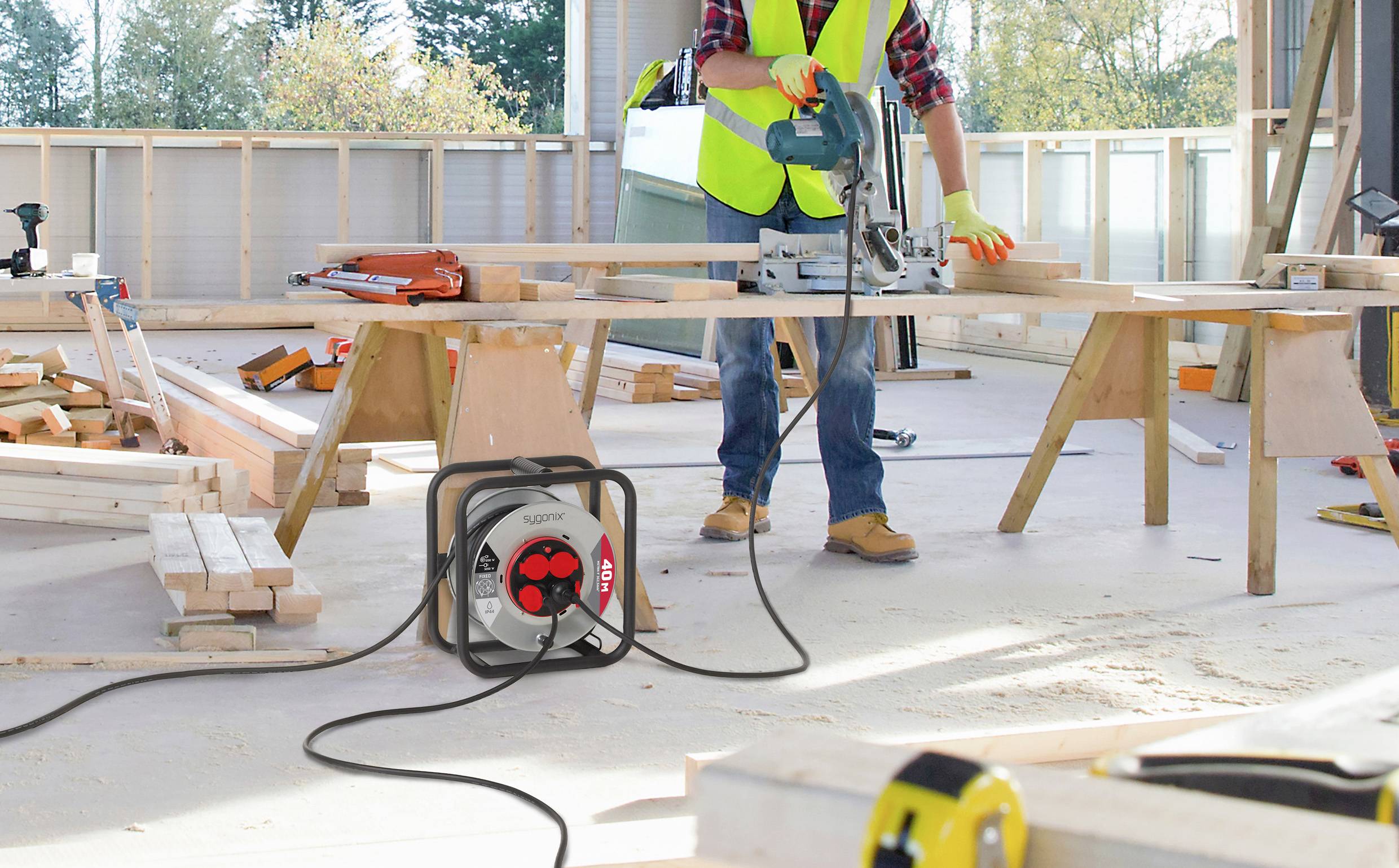 Ein Bauarbeiter benutzt eine Kreissäge auf einem Holzbrett in einem hellen, unfertig wirkenden Gebäude. Werkzeuge und Holzbalken sind verstreut umher.