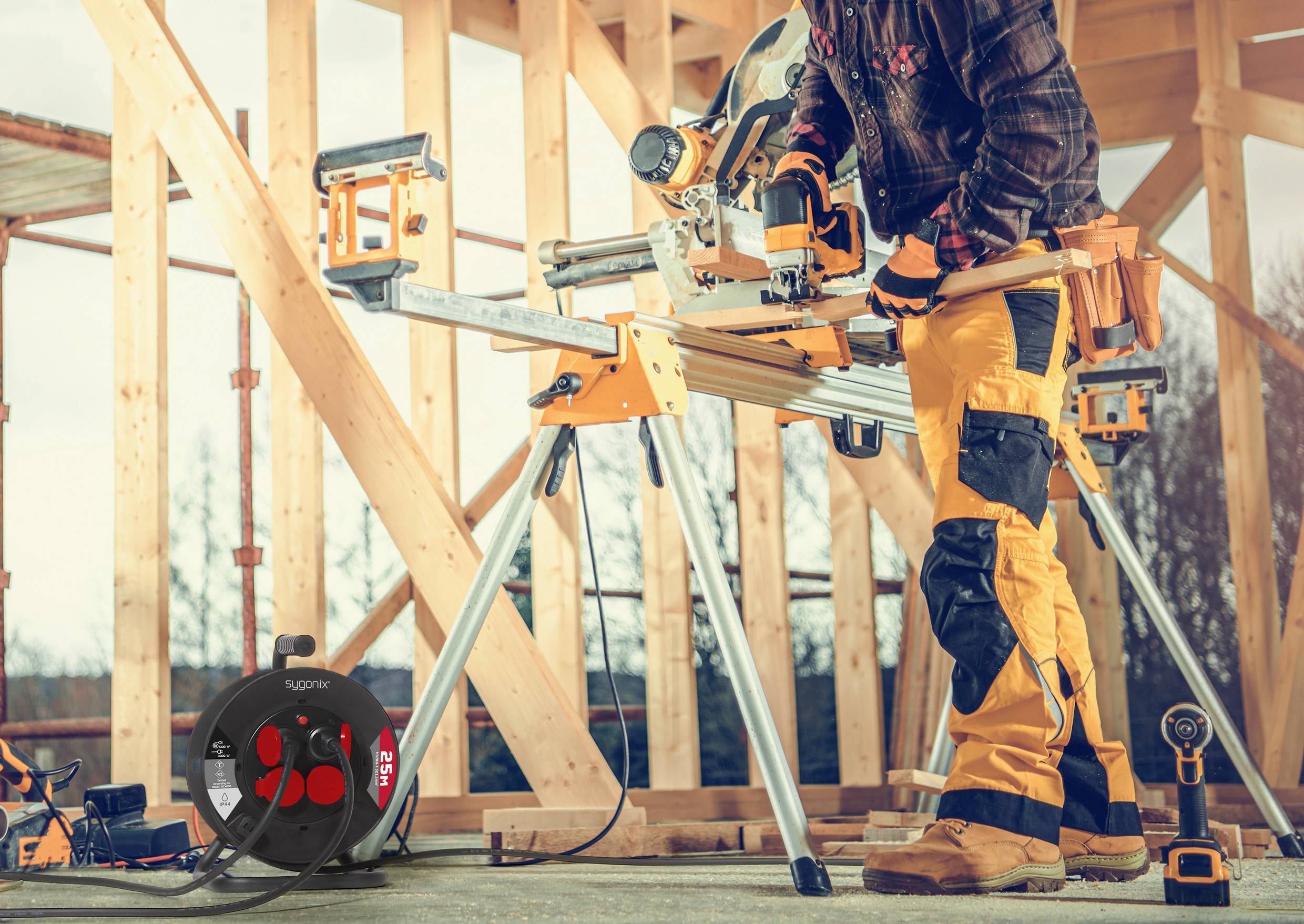 Eine Person in Sicherheitsausrüstung, die mit einer Gehrungssäge auf einer Baustelle arbeitet, umrahmt von unfertig gebauten Holzstrukturen. Ausrüstung in der Nähe.