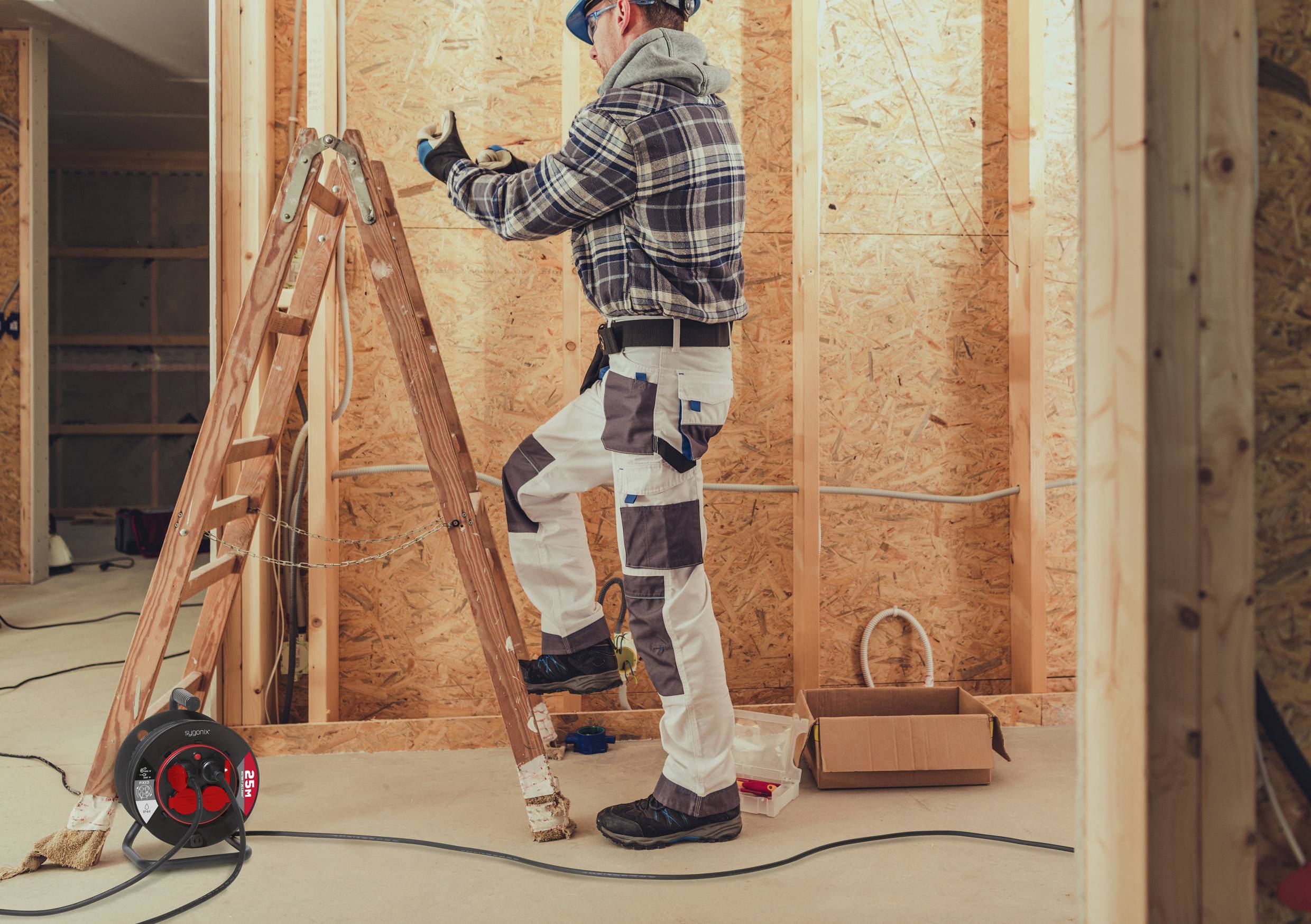 Person auf einer Leiter, der in einem teilweise fertiggestellten Raum arbeitet und Schutzausrüstung trägt, mit einer Verlängerungskabelrolle und einer offenen Kiste in der Nähe.