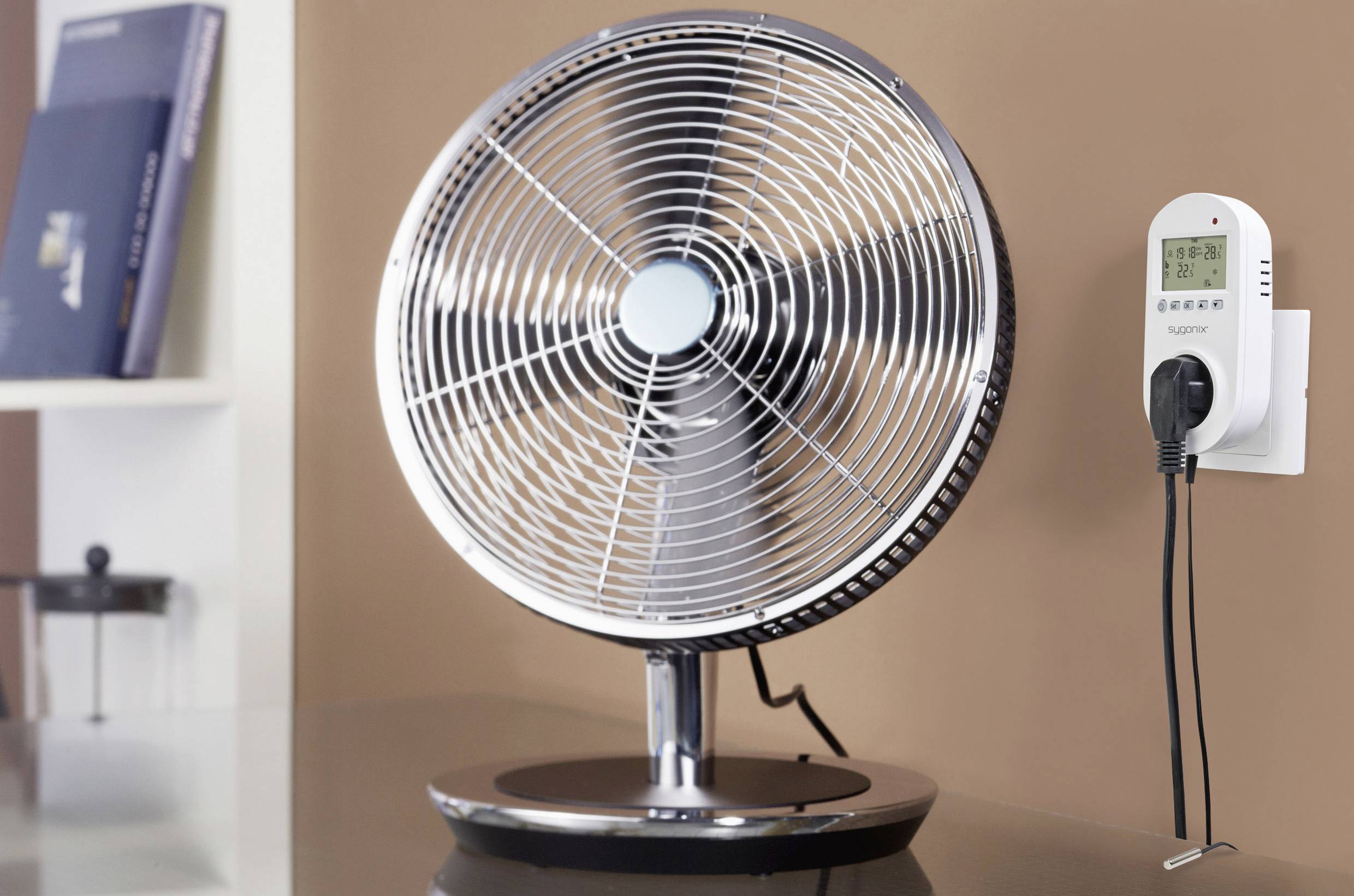 Ein Tischventilator mit Metallgitter steht auf einem Tisch. Rechts daneben ist ein an der Wand montierter Steckdosen-Timer sichtbar.