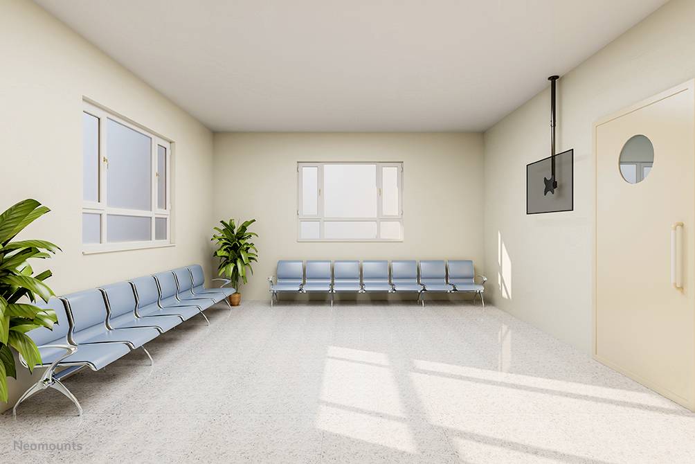 Ein leerer Warteraum mit blauen Stühlen, zwei Pflanzen und einem Fernseher an der Wand. Zwei große Fenster lassen Sonnenlicht herein.