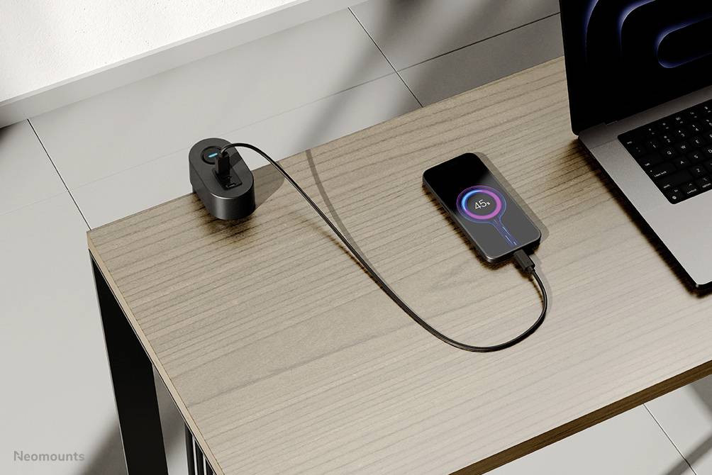 Smartphone auf einem Tisch neben einem Laptop, verbunden über ein Ladekabel zu einer Steckdose. Das Display zeigt 65% Ladestand.