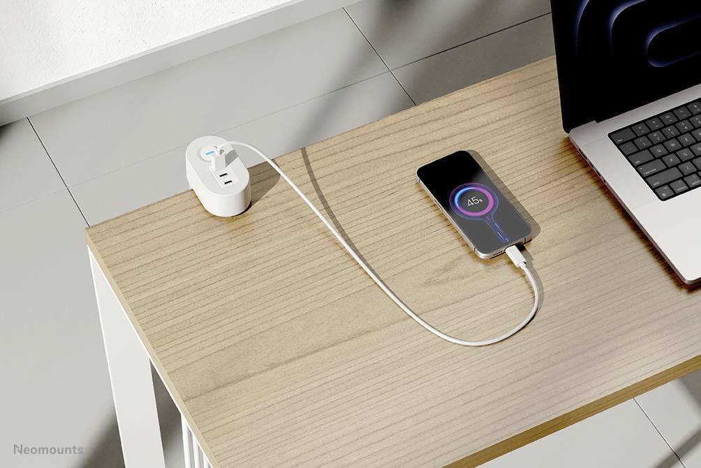 Ein Smartphone lädt auf einem Holztisch neben einem Laptop; es ist mit einem weißen Kabel verbunden, das in eine Steckdose führt.