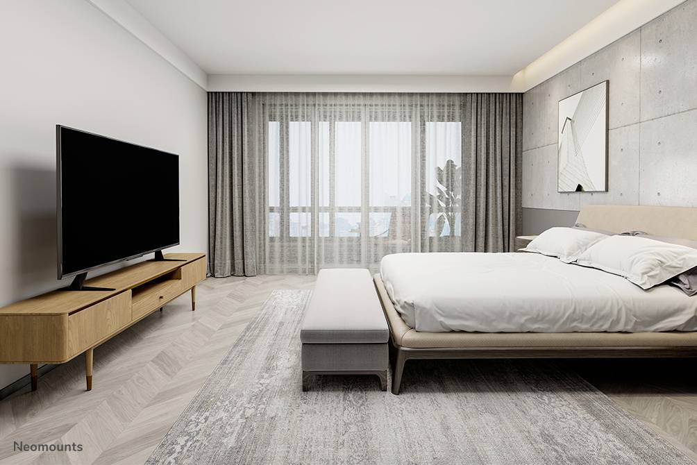 Schlafzimmer mit großem Bett und Flachbildfernseher auf modernem Holzschrank. Große Fenster mit grauen Vorhängen, helle Wände.