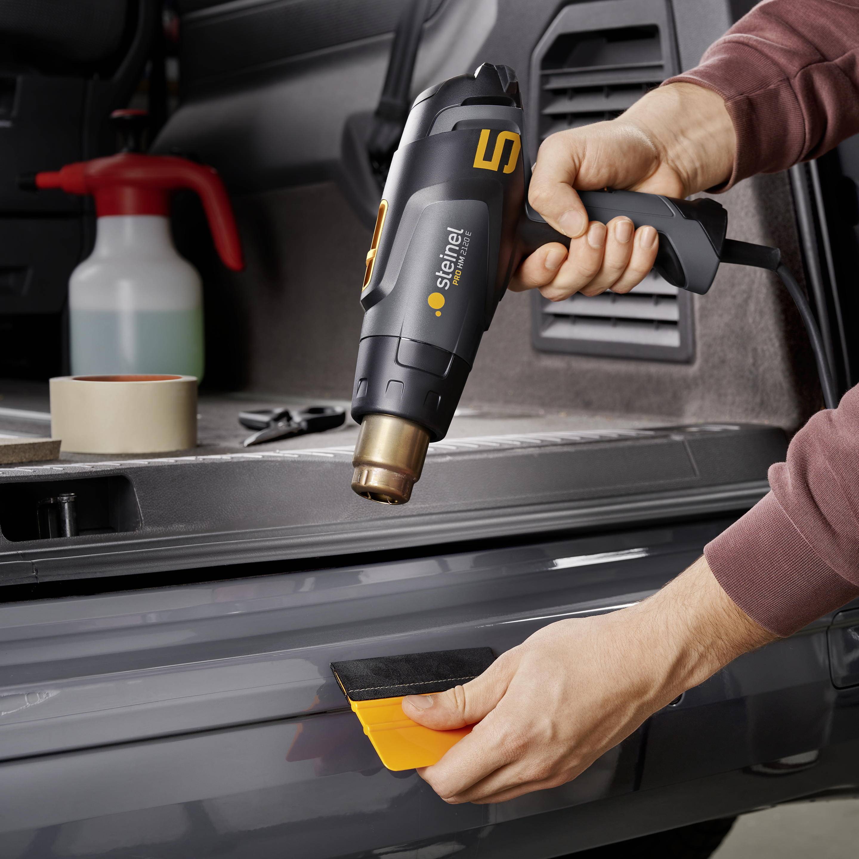 Eine Person verwendet ein Heißluftgebläse, um eine Folie auf eine Auto-Stoßstange aufzutragen. Im Hintergrund sind Werkzeuge zu sehen.
