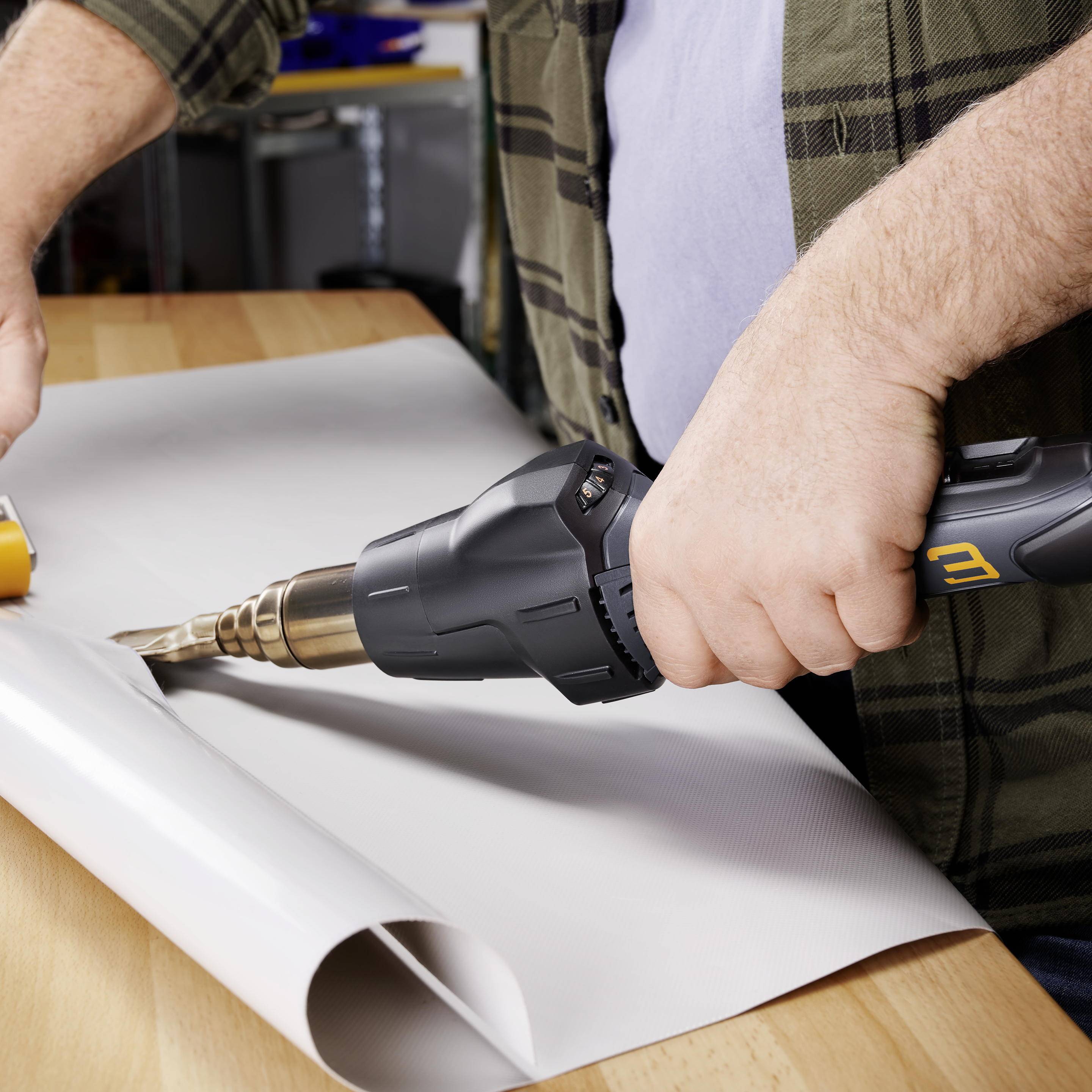 Eine Person glättet mit einer Heißluftpistole weiße Folie auf einem Holztisch. Werkzeug im Hintergrund.