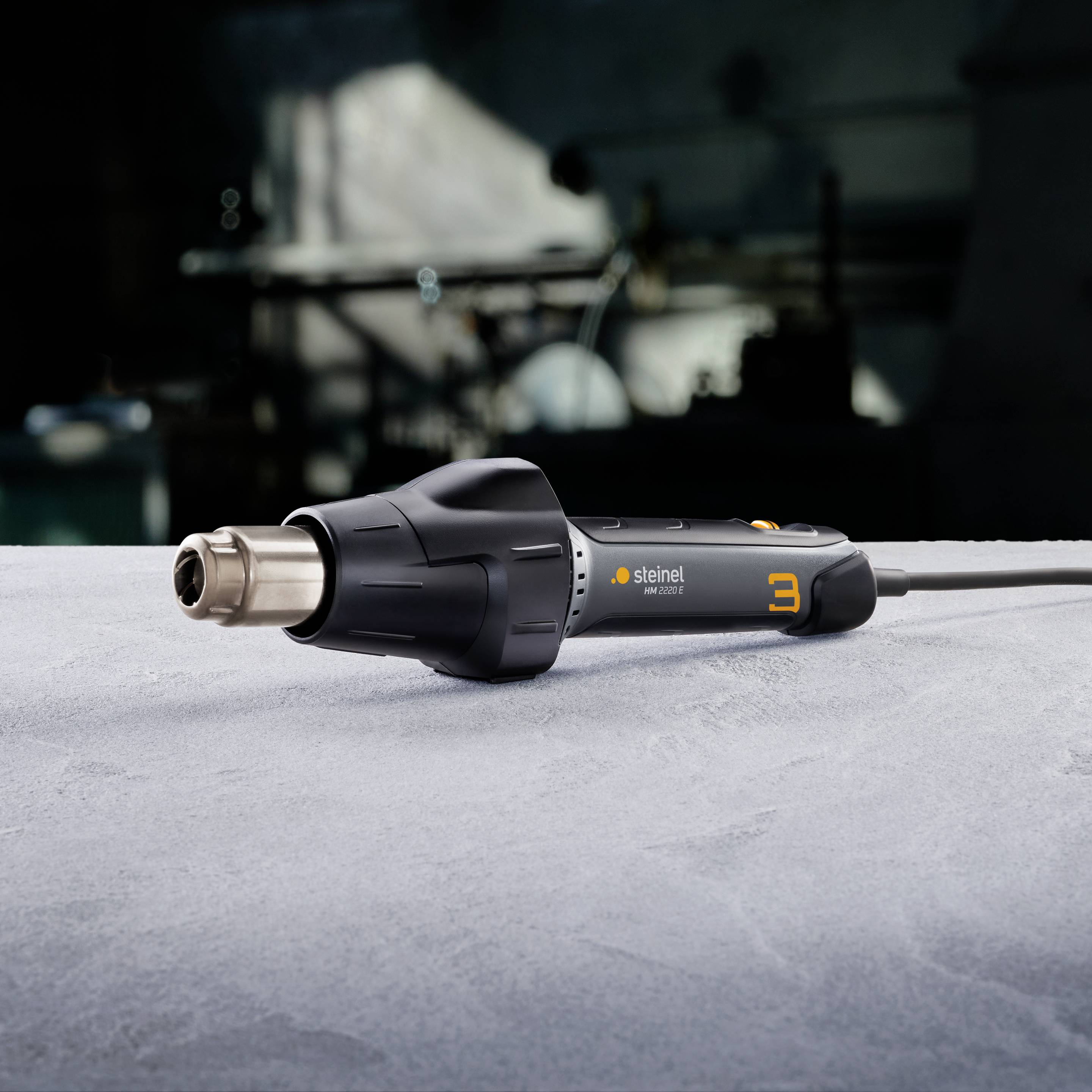 Ein schwarzer, elektrischer Heißluftfön mit digitaler Anzeige steht auf einem grauen Tisch vor einem unscharfen Werkstatt-Hintergrund.