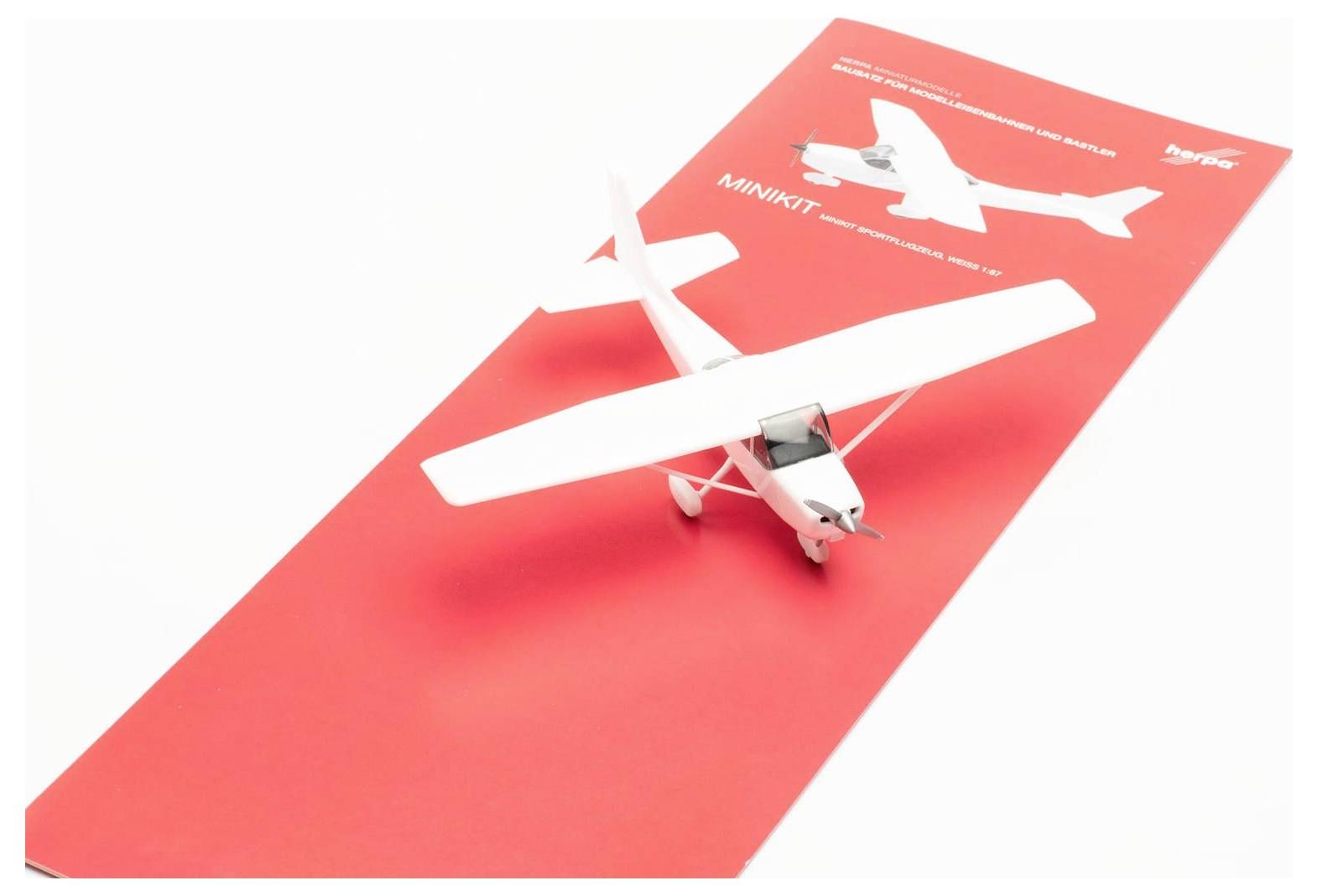 Ein weißes Modellflugzeug auf einer roten Verpackung mit der Aufschrift 'Minikit'. Das Flugzeug steht diagonal, Vorderseite zeigt nach links.
