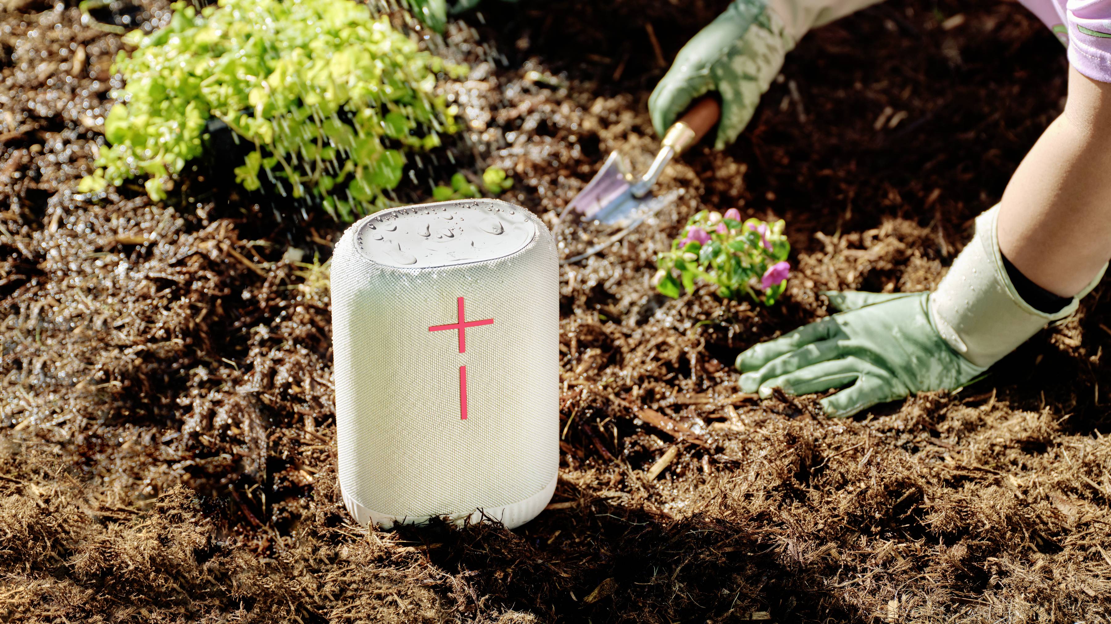 Eine Person pflanzt Blumen in einem Garten. Im Vordergrund steht ein Lautsprecher auf der Erde.