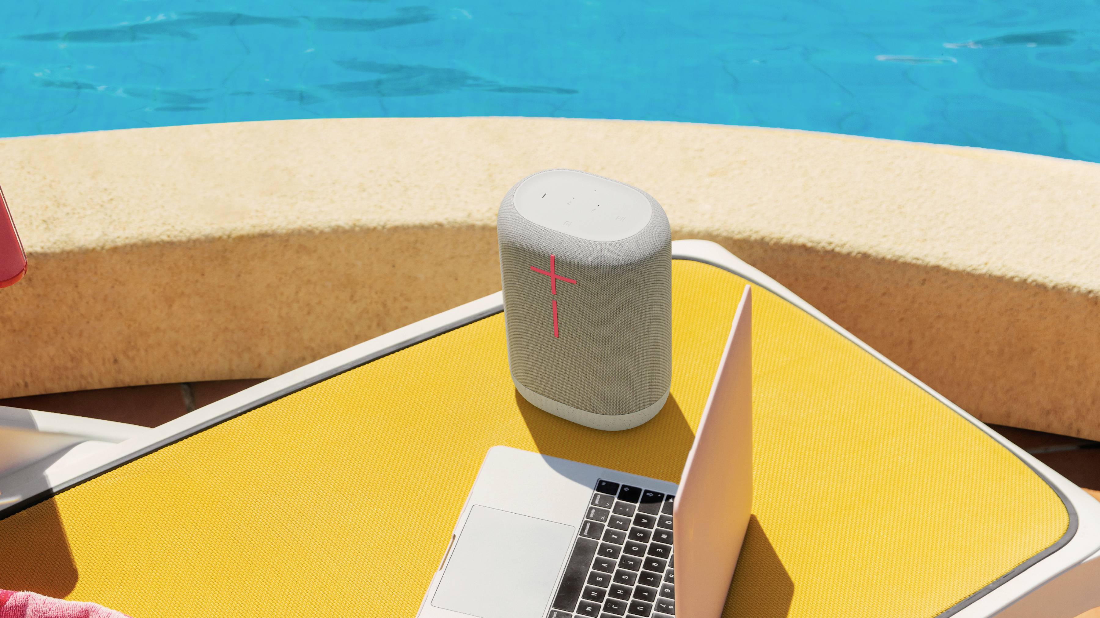 Ein Laptop und ein Lautsprecher stehen auf einer gelben Liege neben einem Pool. Der Lautsprecher hat ein leuchtendes rotes Pluszeichen.