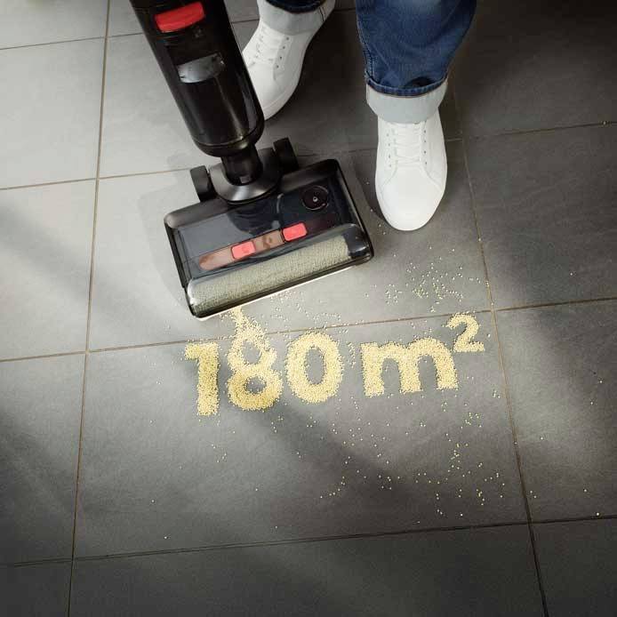 Staubsauger entfernt Krümel von schwarzem Fliesenboden. Text auf Boden: '180 m²'. Zwei weiße Schuhe sichtbar.