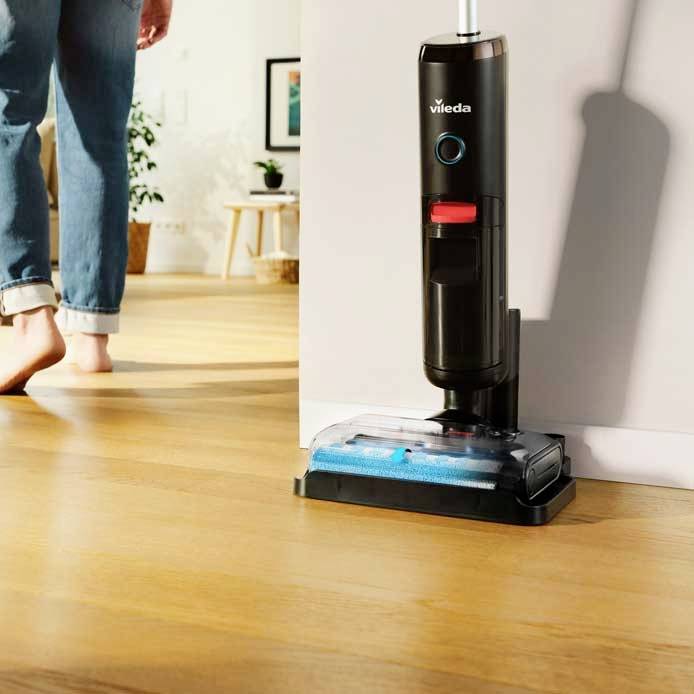 Ein Mensch geht barfuß auf einem Holzfußboden, während ein stehender Bodenreiniger der Marke Vileda sichtbar im Raum steht.