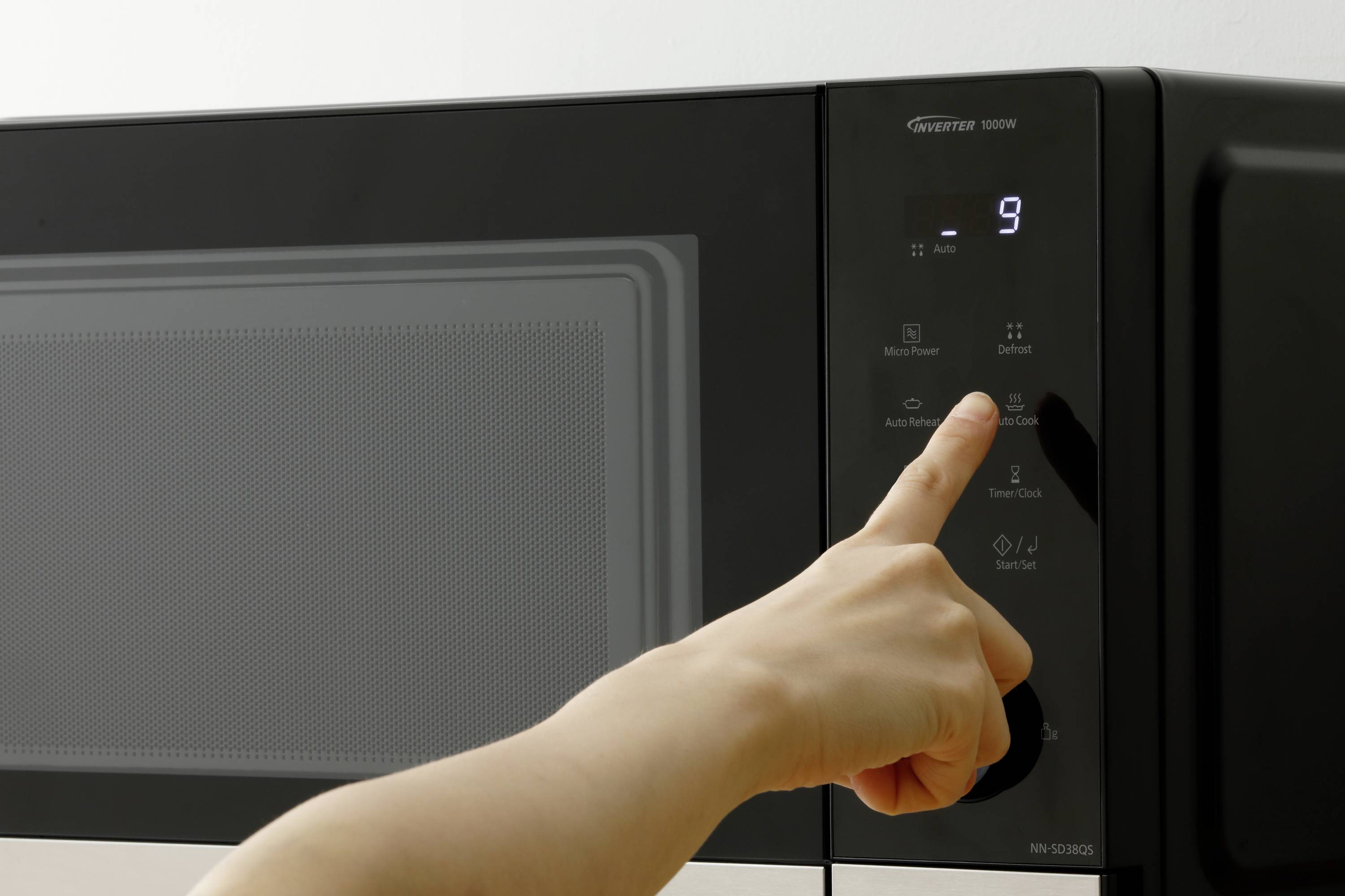 Eine Hand drückt die Starttaste einer schwarzen Mikrowelle, wobei auf dem Display die Zahl 9 leuchtet, was die eingestellte Zeit anzeigt.