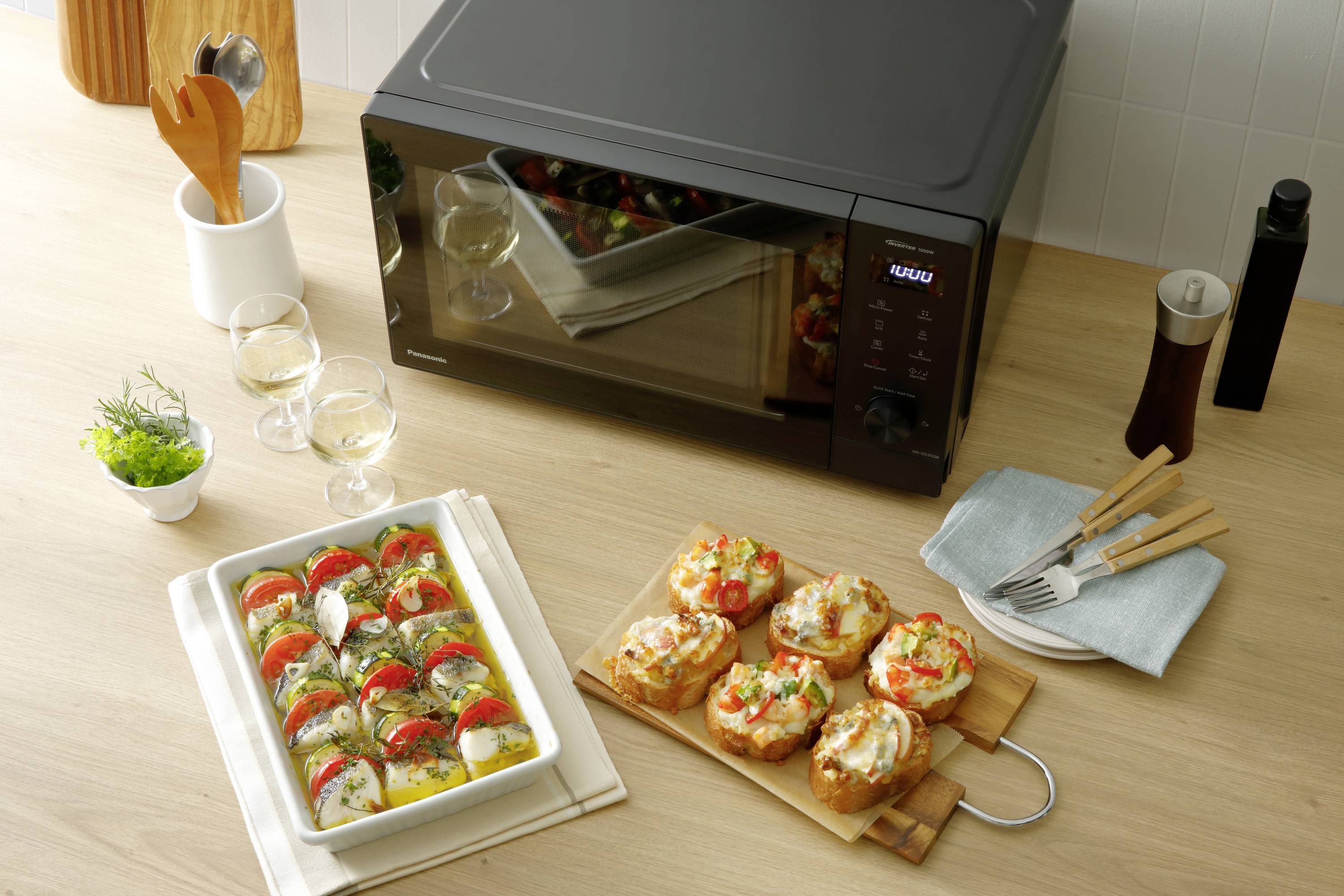 Mikrowelle auf Küchentresen mit einem gebackenen Auflauf und belegten Brötchen. Daneben stehen zwei Weingläser und Küchenutensilien.