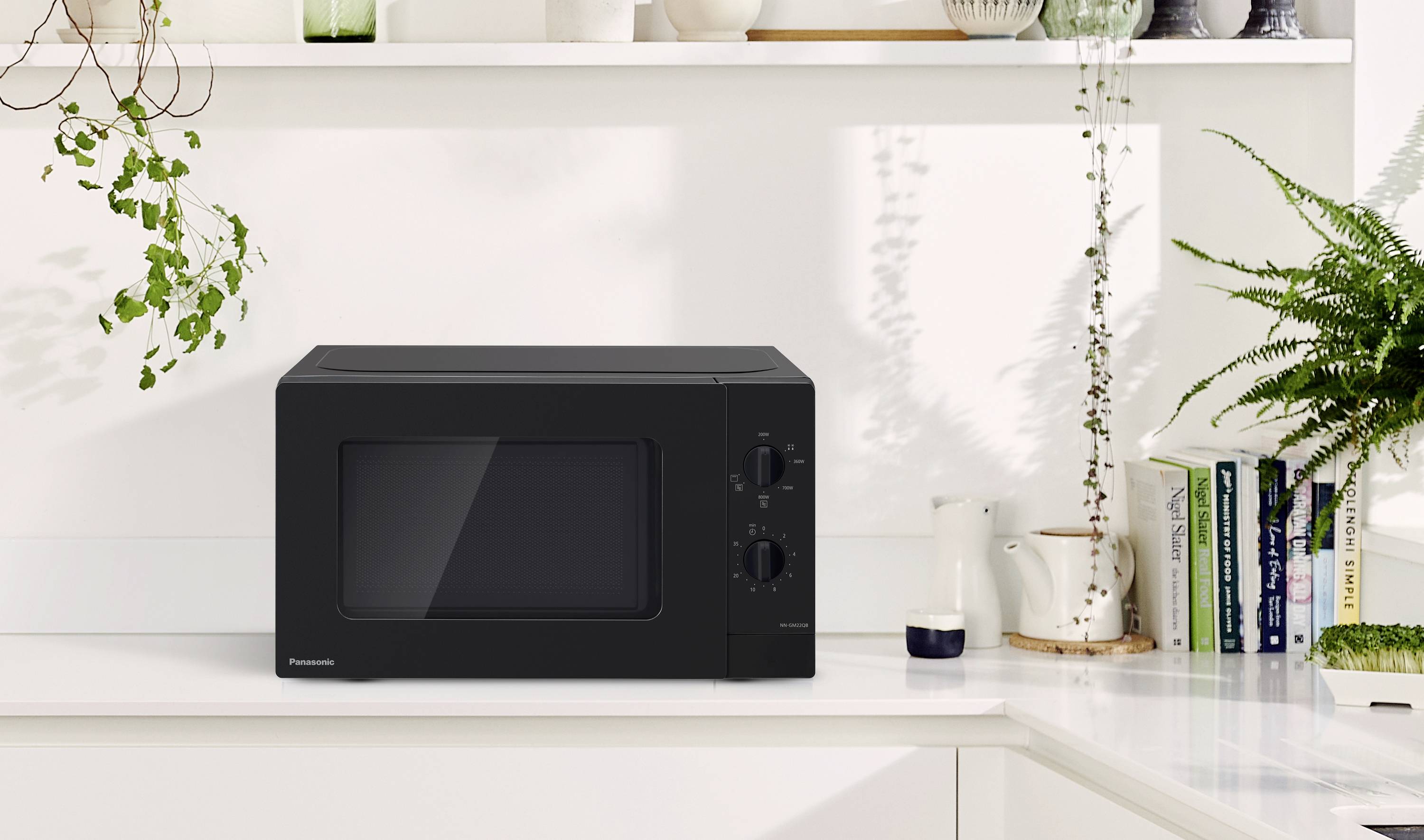 Schwarze Mikrowelle auf weißer Küchenarbeitsplatte, umgeben von Pflanzen und Geschirr. Regale mit Büchern im Hintergrund.