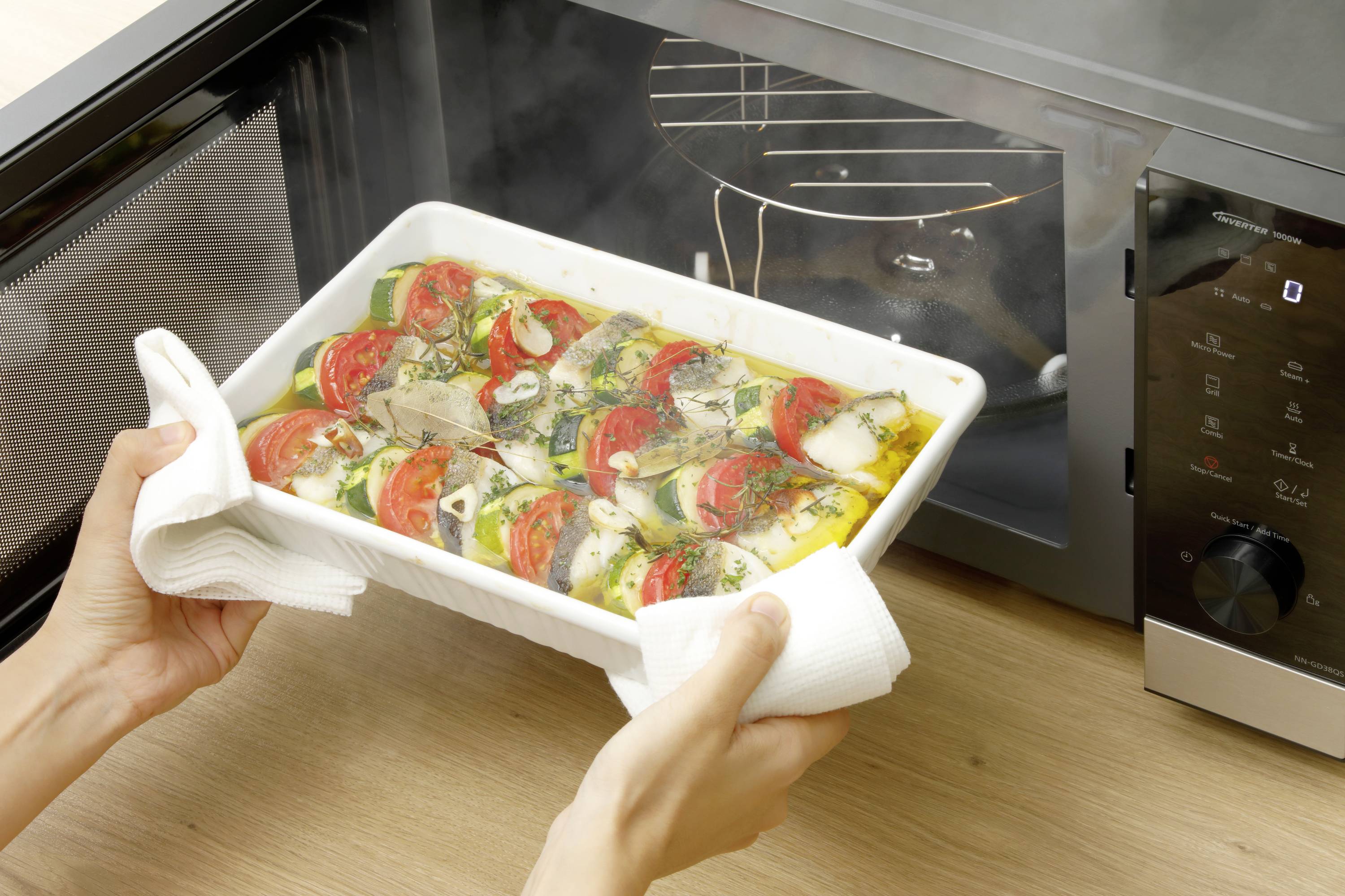 Eine Person hält ein gerolltes Gemüsegericht in einer rechteckigen weißen Form, bereit, es in eine graue Mikrowelle zu stellen.