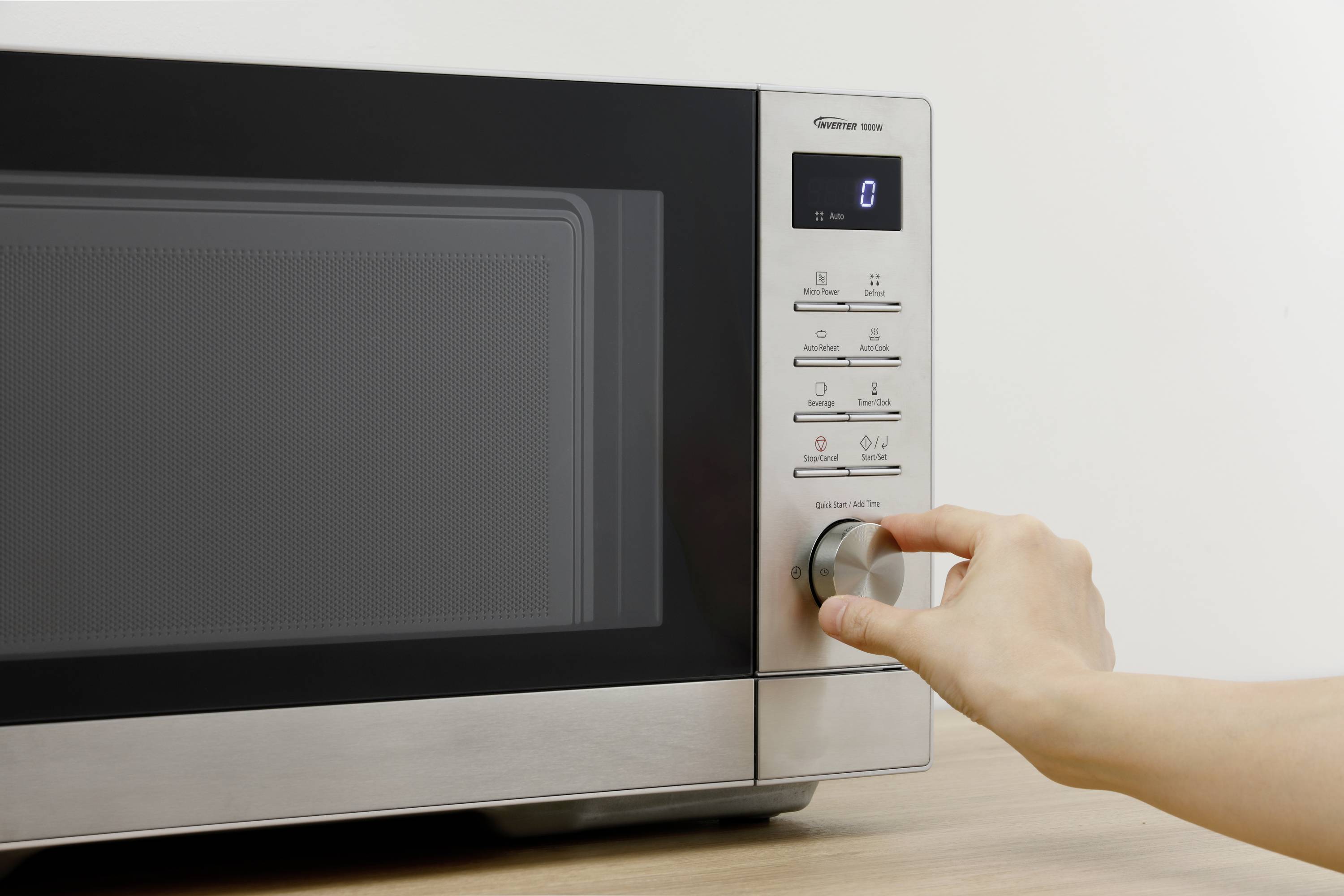 Eine Hand dreht den Einstellknopf einer modernen Mikrowelle. Das Display zeigt die Uhrzeit. Rechteckiges Design, Edelstahloberfläche.