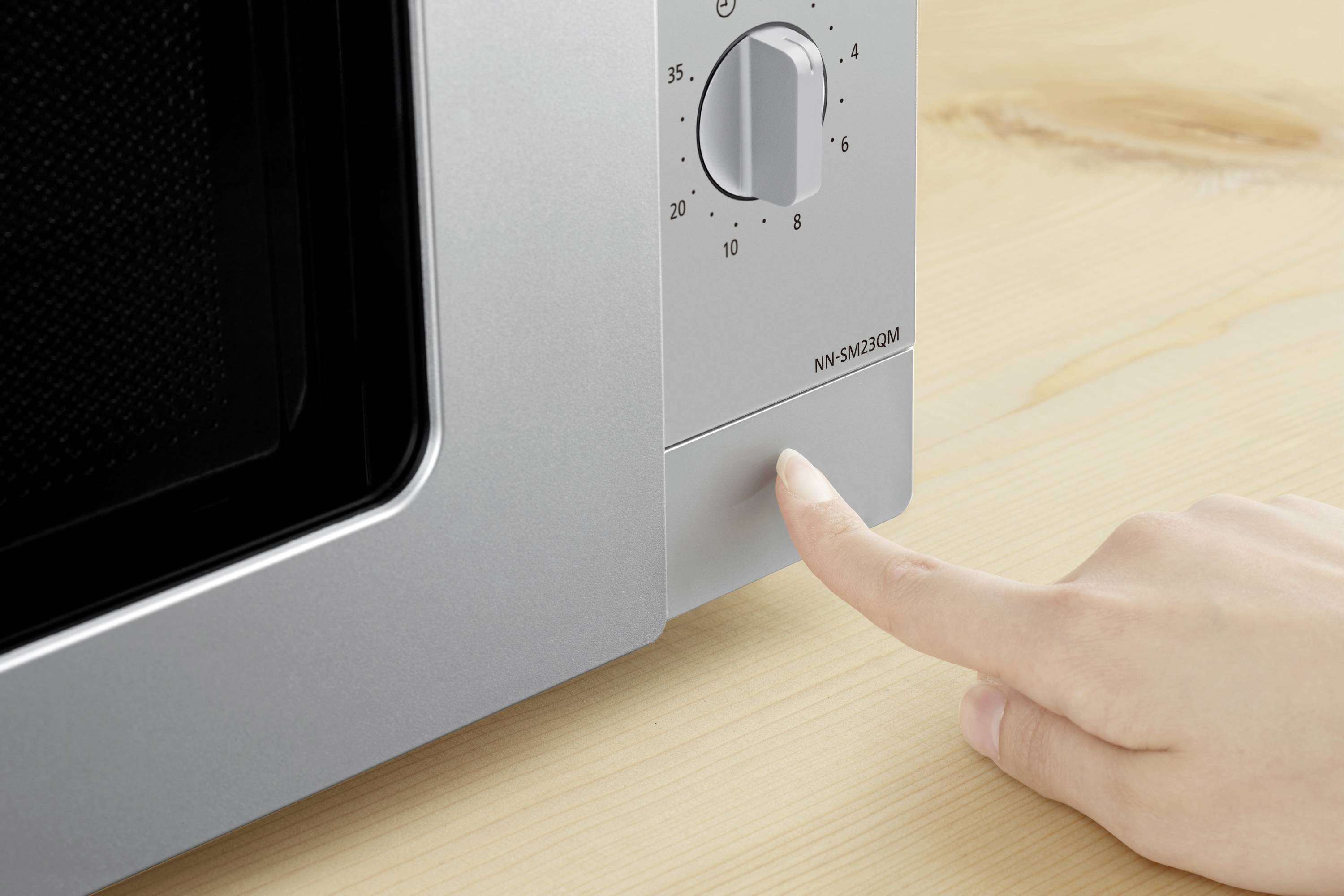 Eine Hand drückt die Taste an einem silbernen Mikrowellenherd, der auf einem Holztisch steht.
