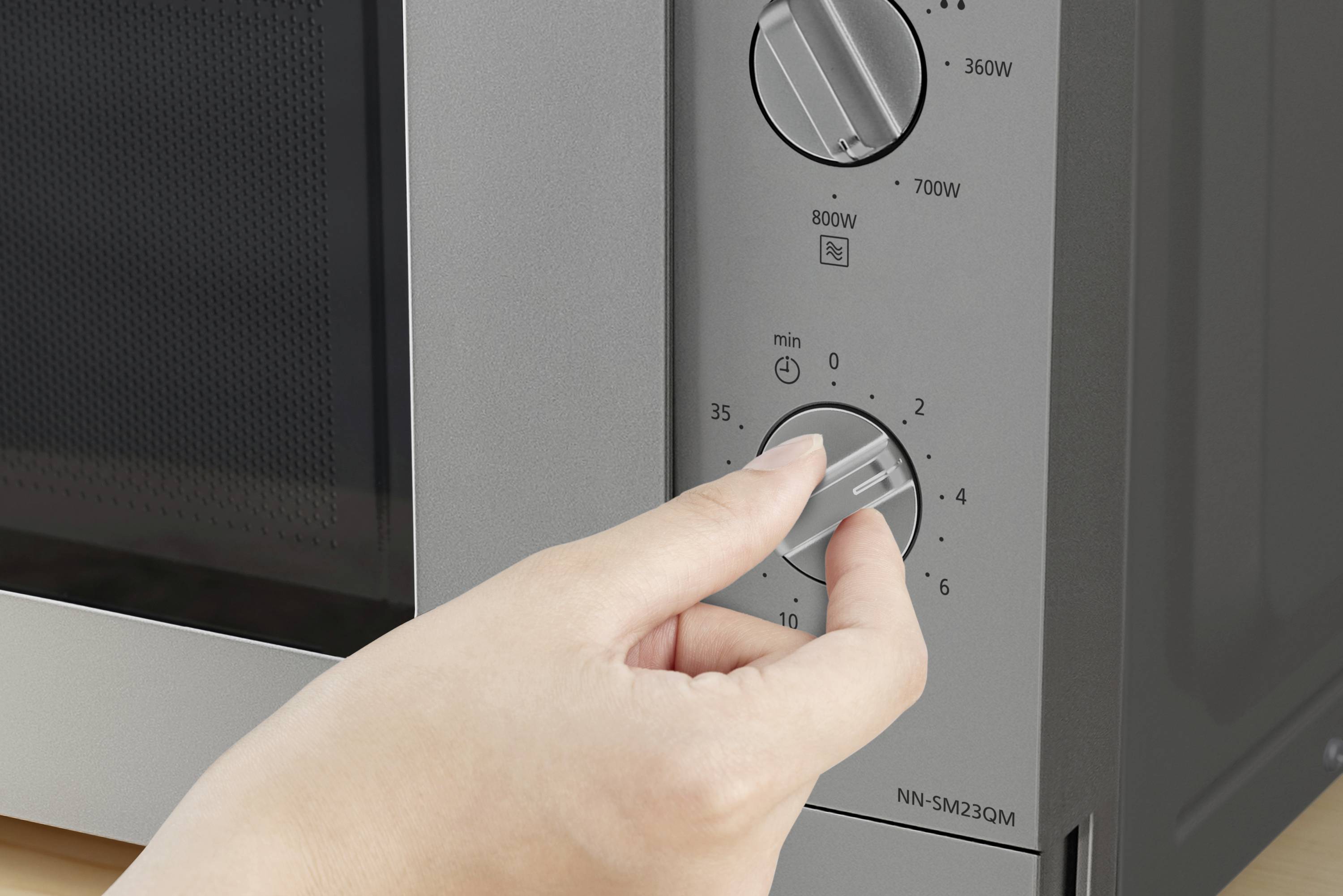 Eine Hand dreht den Timer-Drehknopf einer Mikrowelle, die in Minuten eingestellt wird. Zwei weitere Drehknöpfe sind für die Leistung.