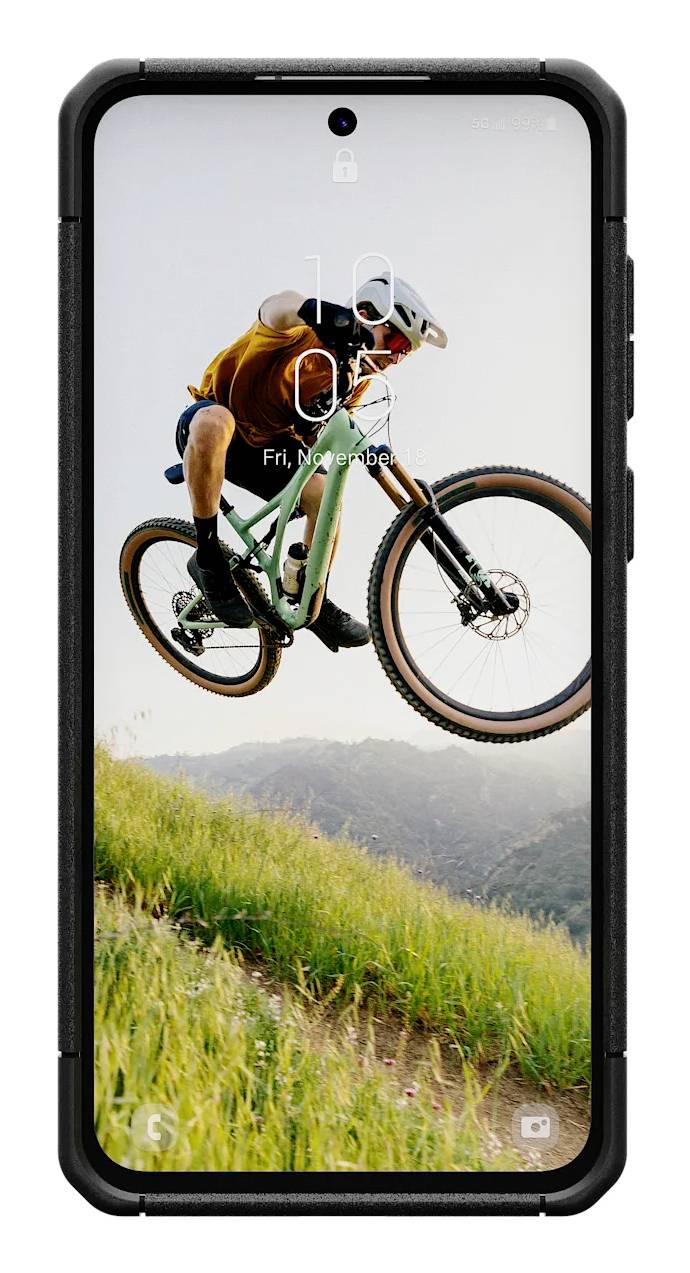 Fahrradfahrer in der Luft, trägt Helm und orange Kleidung, fährt über grasbewachsene Hügel. Auf dem Smartphone-Bildschirm ist die Uhrzeit sichtbar.