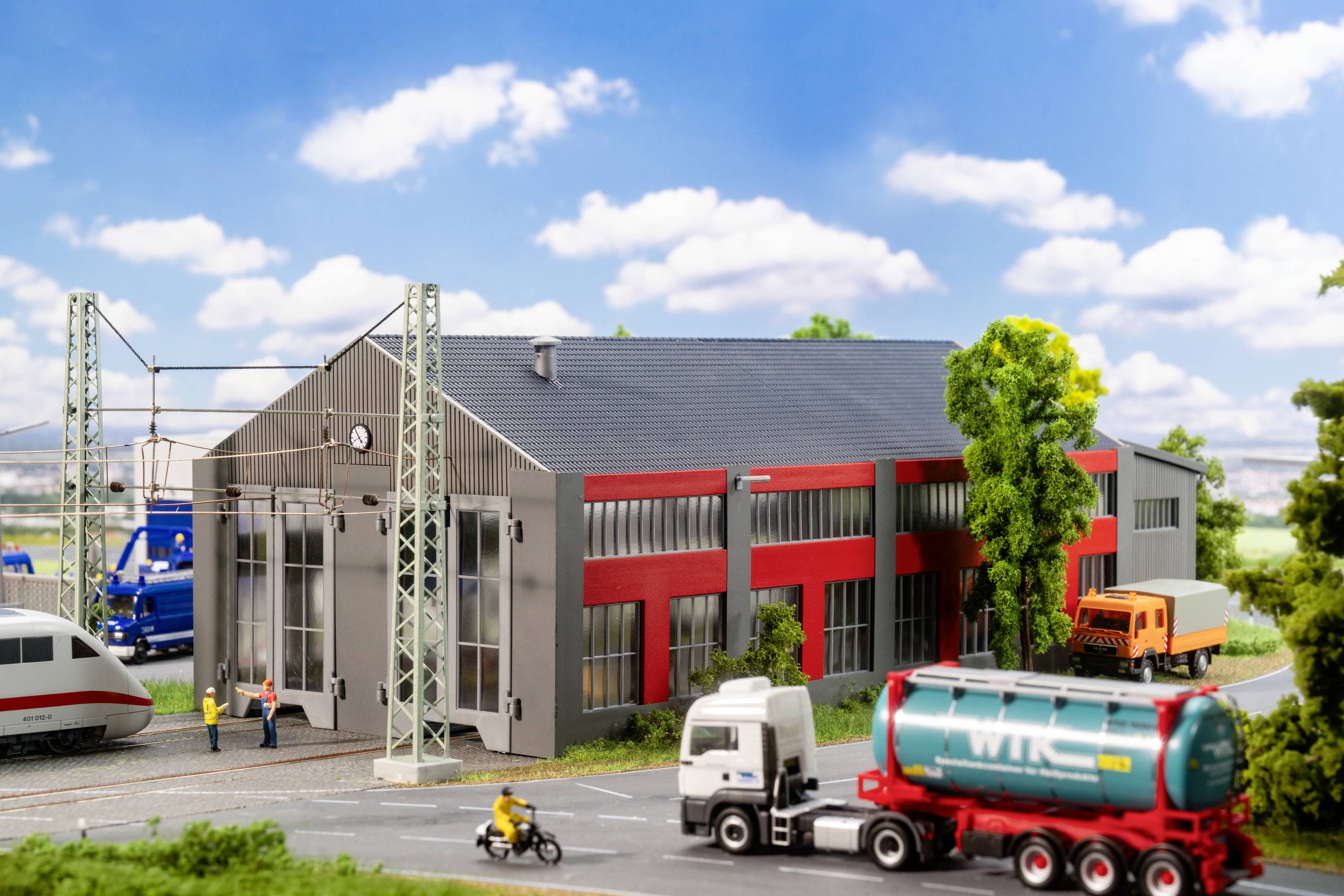 Miniatur-Landschaft mit einem Bahndepot, LKWs, einem Zug und Figuren auf einer Straße. Gebäude im Hintergrund mit roter Fassade.