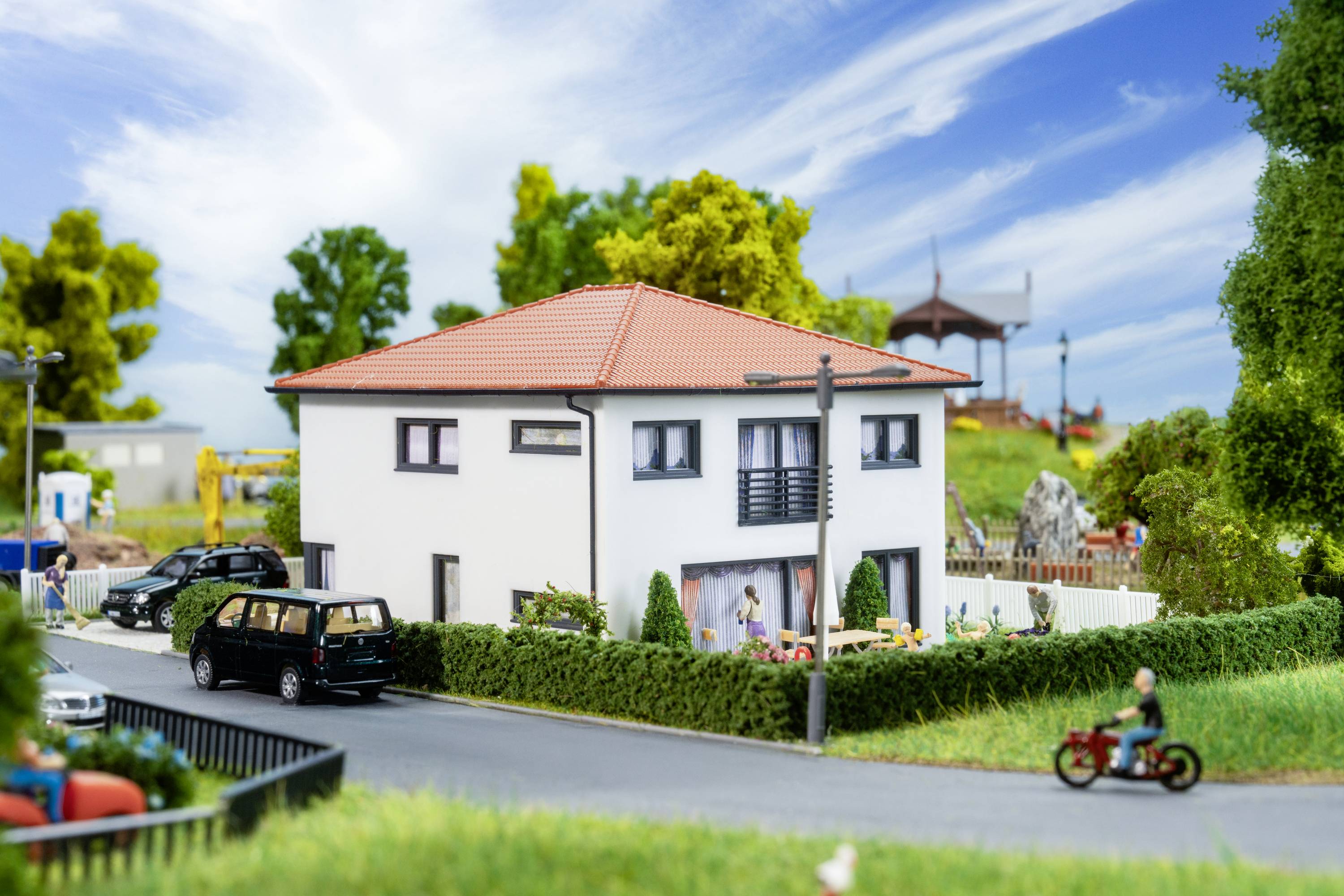 Ein modernes zweistöckiges weißes Haus mit rotem Dach steht an einer Straße, umgeben von Bäumen und einem Garten. Im Hintergrund ist ein Pavillon sichtbar.