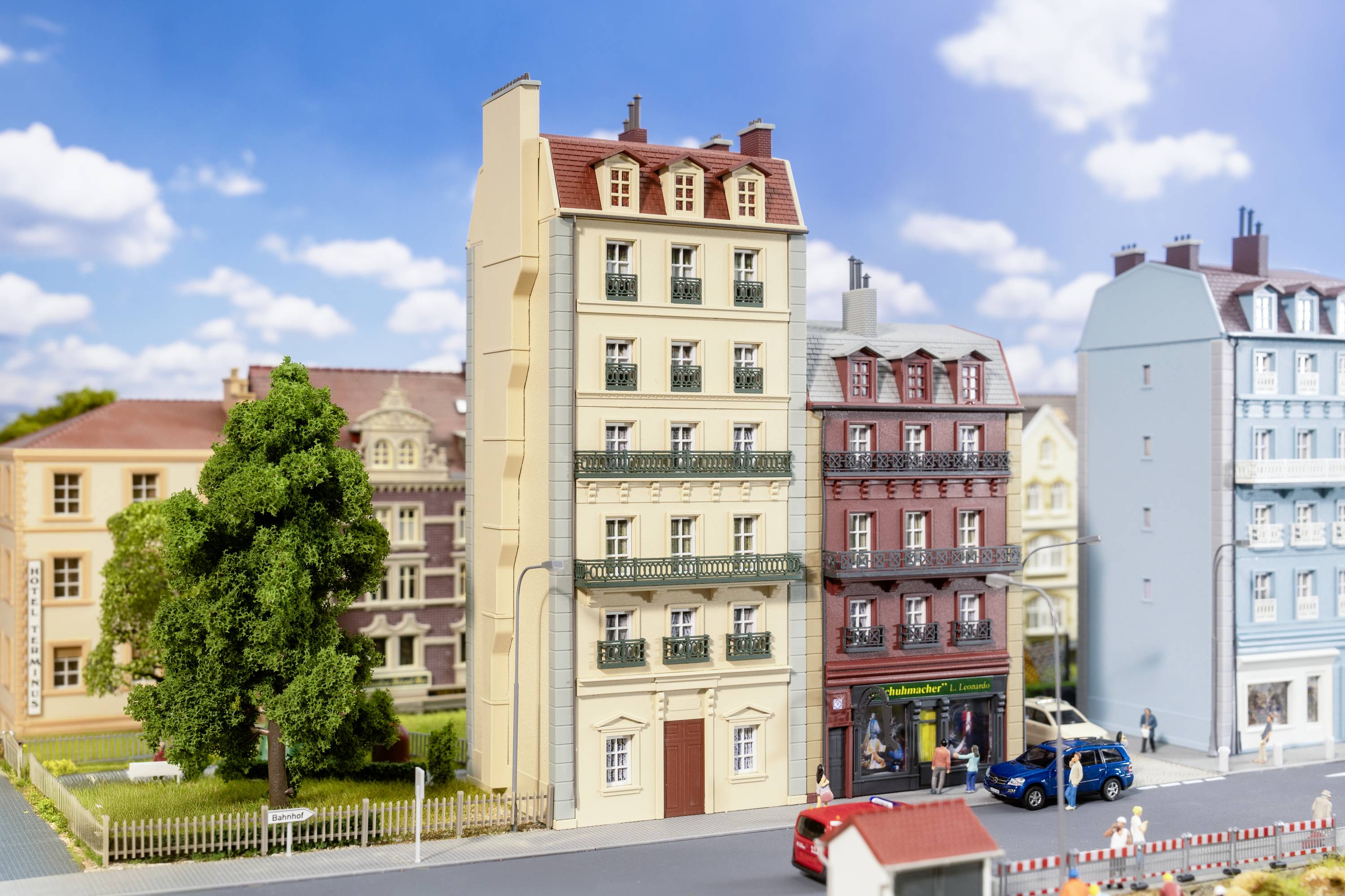 Miniaturstadt-Szene mit mehreren detailreichen Gebäuden, Autos und Fußgängern auf einer Straße unter einem blauen Himmel mit Wolken.