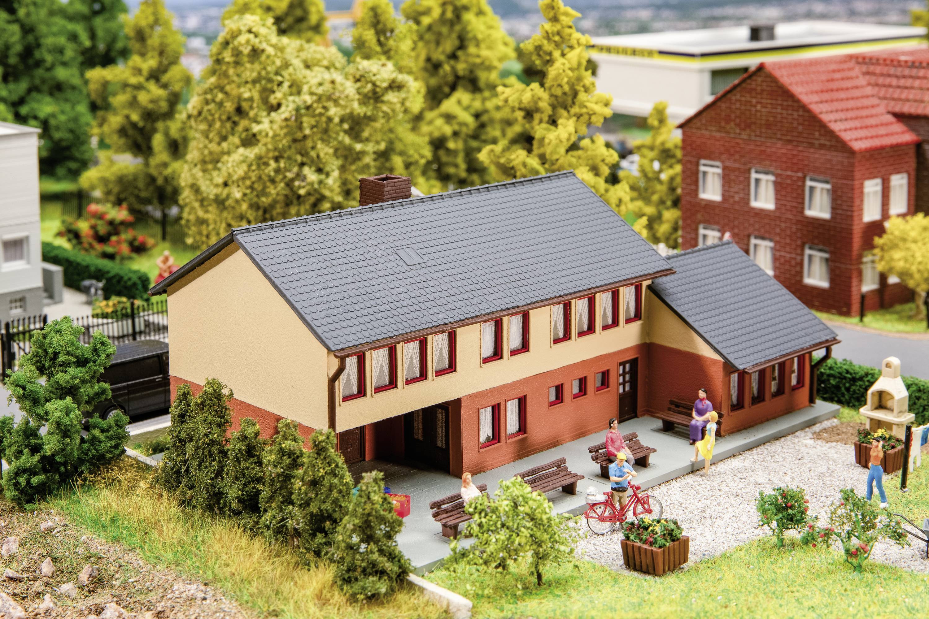 Ein Miniaturmodell zeigt ein Gebäude mit rotem Ziegeldach, umgeben von Bäumen und Figuren, in einer Dorfkulisse.