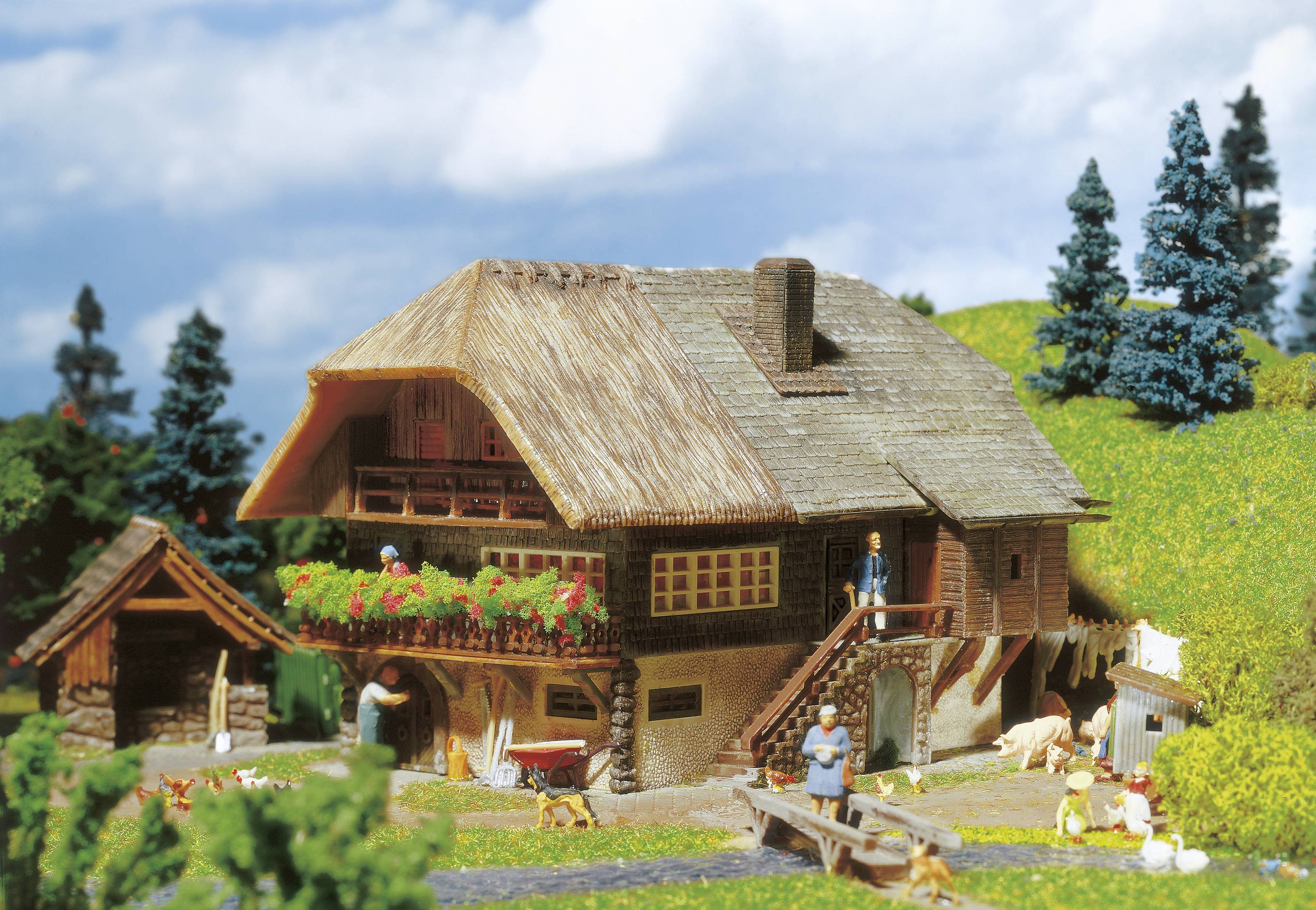 Ein Miniatur-Bauernhaus mit Strohdach, umgeben von Figuren, Hunden und Tieren auf einem Grasfeld mit Bäumen im Hintergrund.