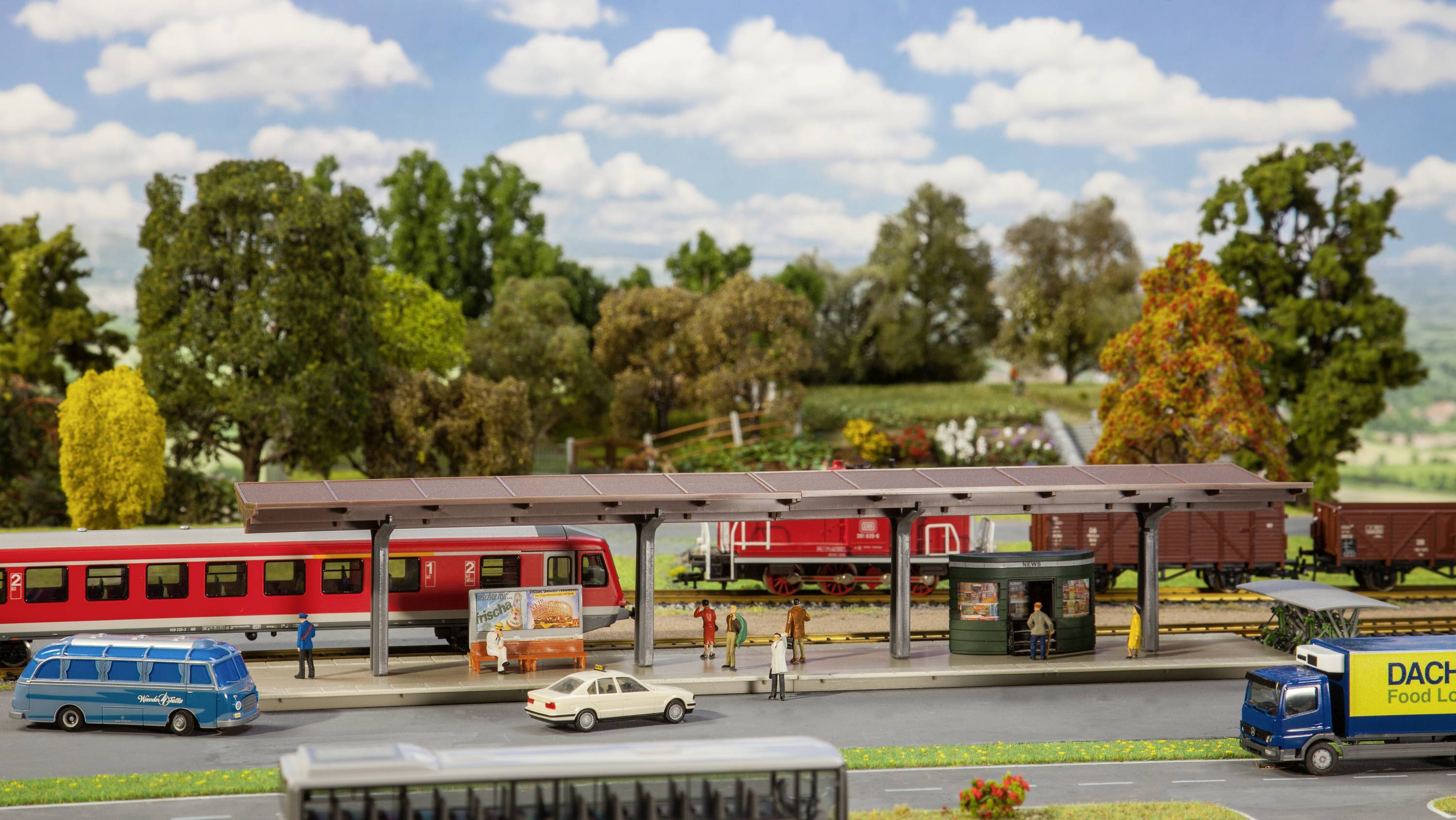 Ein Miniaturbahnhof mit rotem Zug, umgeben von Bäumen und Straßen, zeigt Menschen beim Ein- und Aussteigen.
