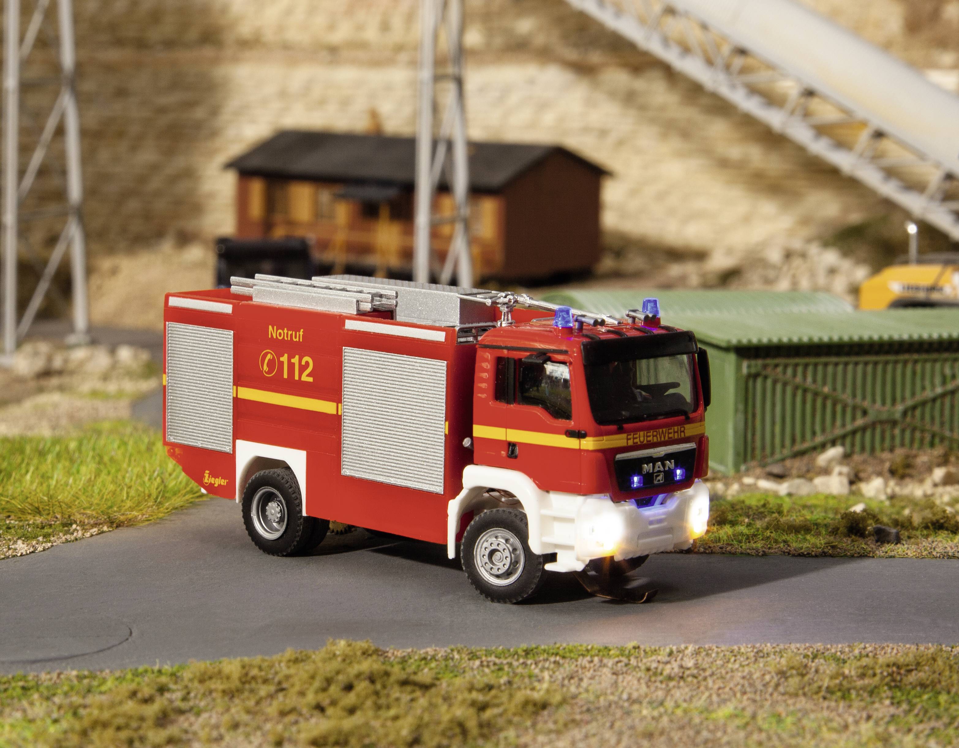 Ein rotes Modell-Feuerwehrauto mit der Aufschrift 'Notruf 112' fährt auf einer Miniaturlandschaftsstraße, Gebäude im Hintergrund.