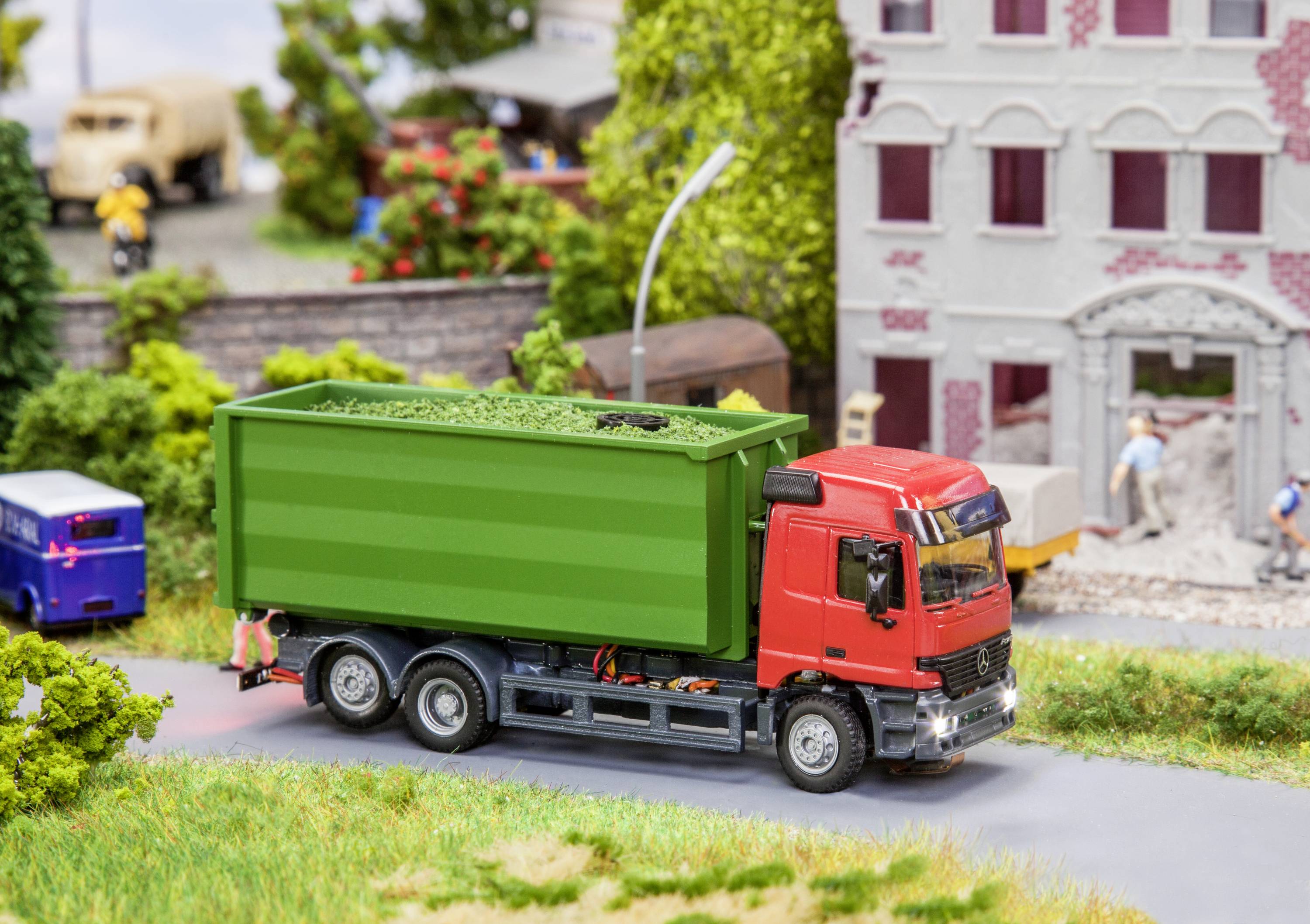 Ein rotes Modell-Lkw mit einem grünen Container fährt auf einer Miniatur-Straße neben einem Gebäude und kleinen Figuren.