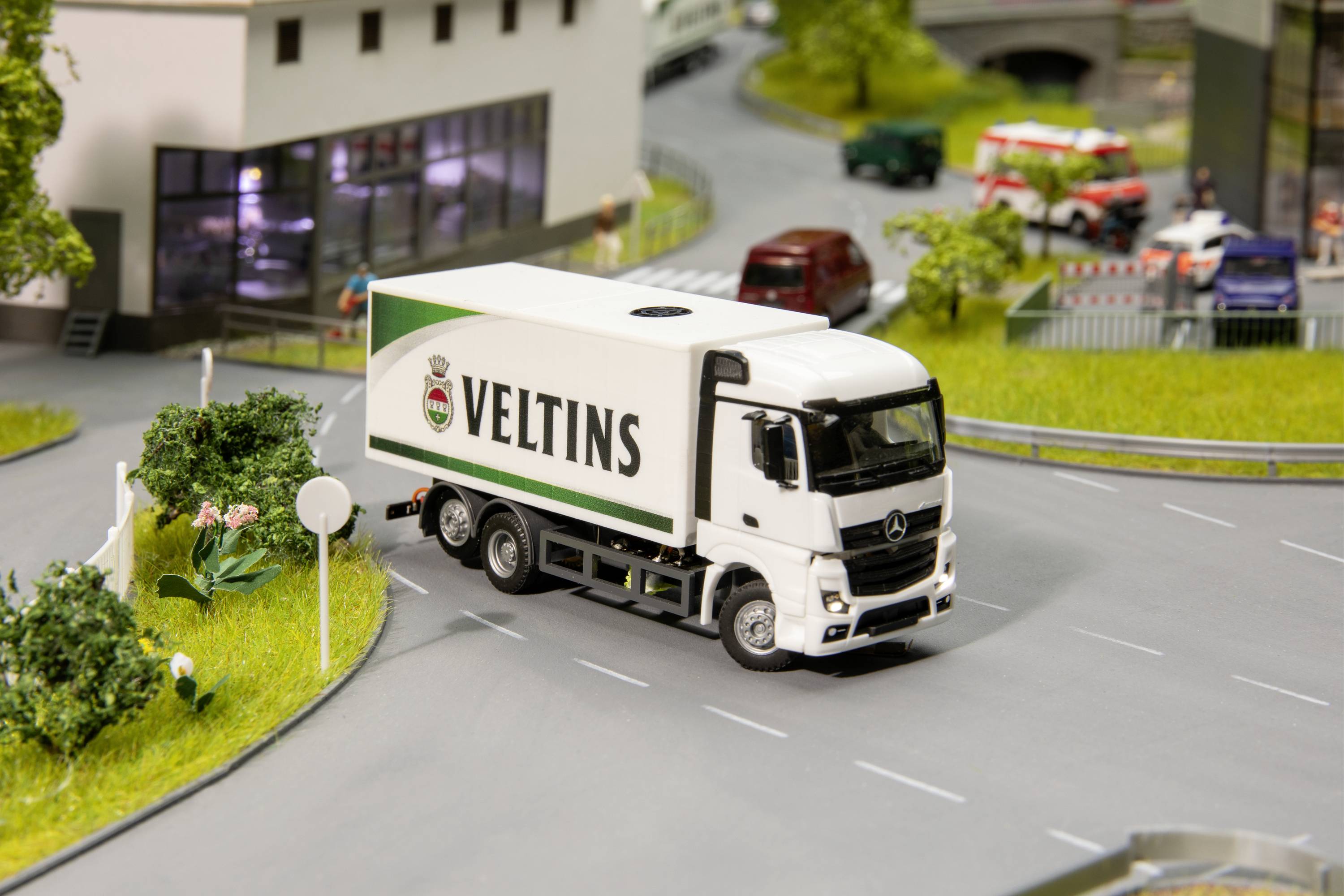 Ein weißer Lastwagen mit der Aufschrift 'Veltins' fährt auf einer modellierten Straßenlandschaft. Im Hintergrund sind Gebäude und Bäume zu sehen.