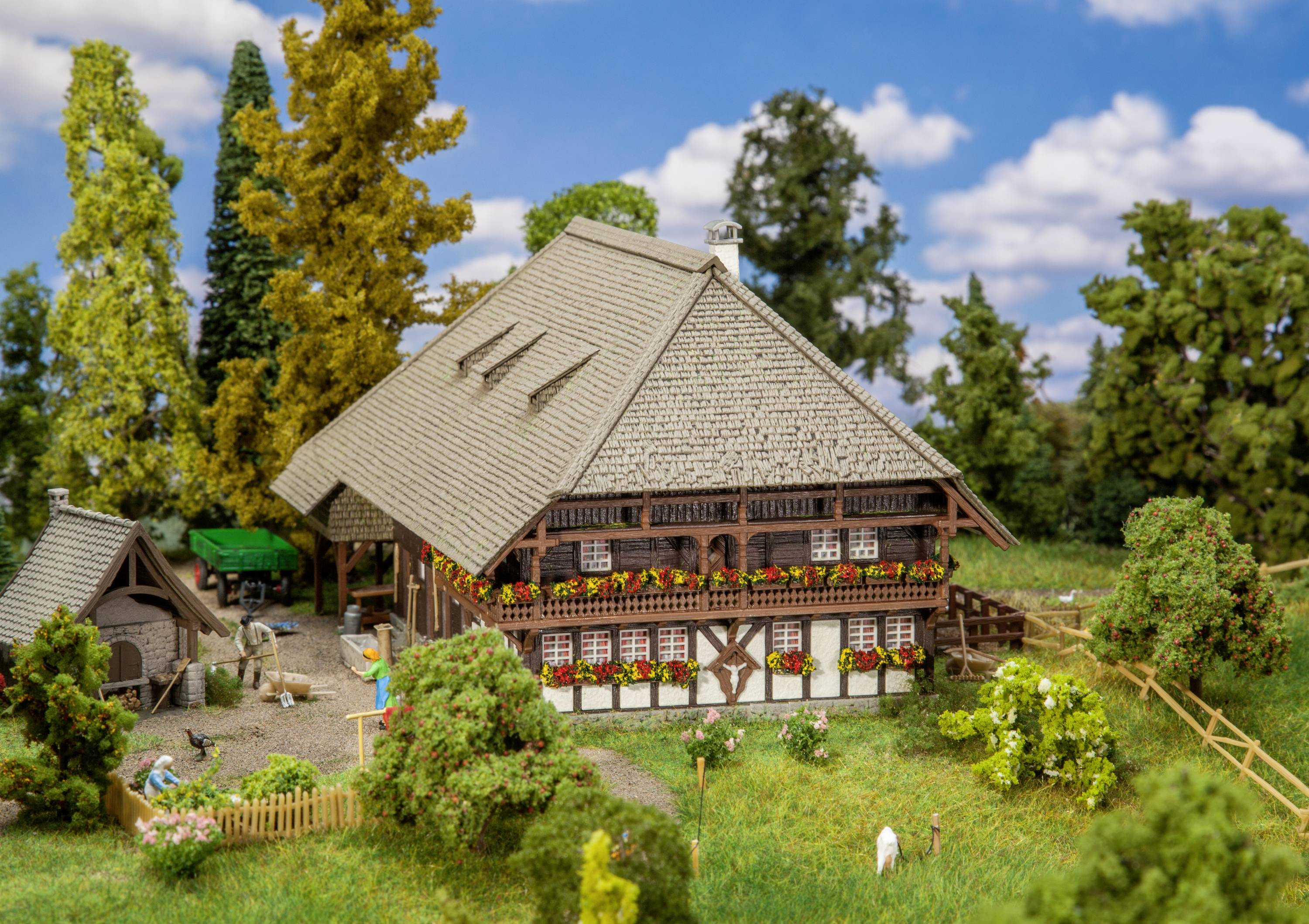 Traditionelles Fachwerkhaus in ländlicher Umgebung mit Blumenschmuck an Balkonen, umgeben von Bäumen und einem kleinen Hof im Vordergrund.
