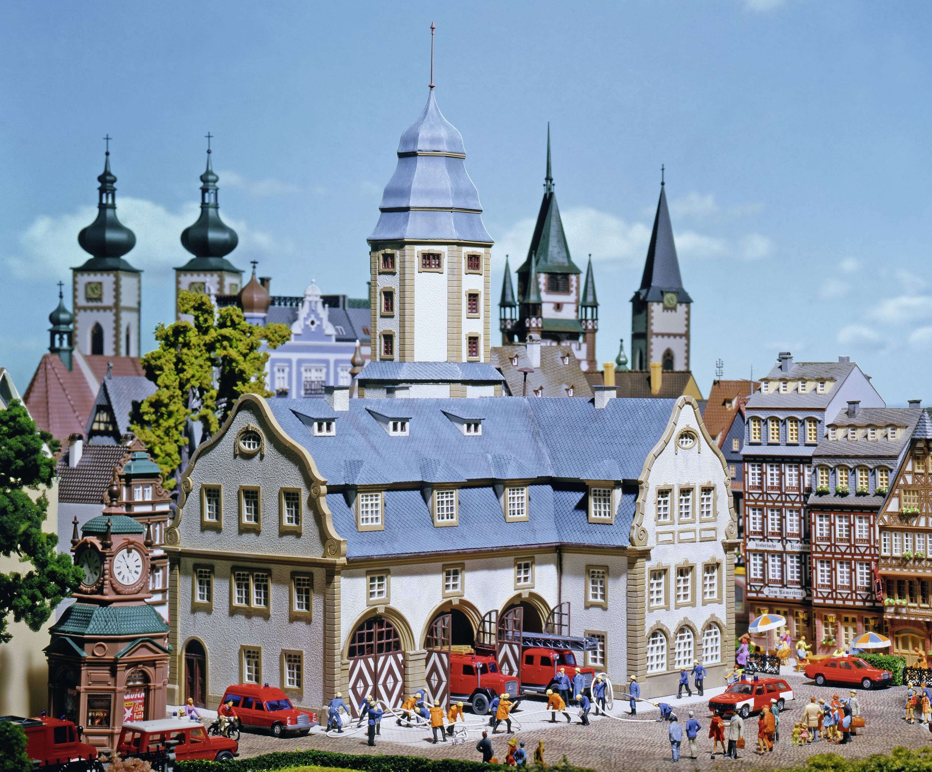 Modellstadt mit historischen Gebäuden, Kirchtürmen und kleinen Figuren, die auf dem Marktplatz spazieren. Autos parken in der Nähe.