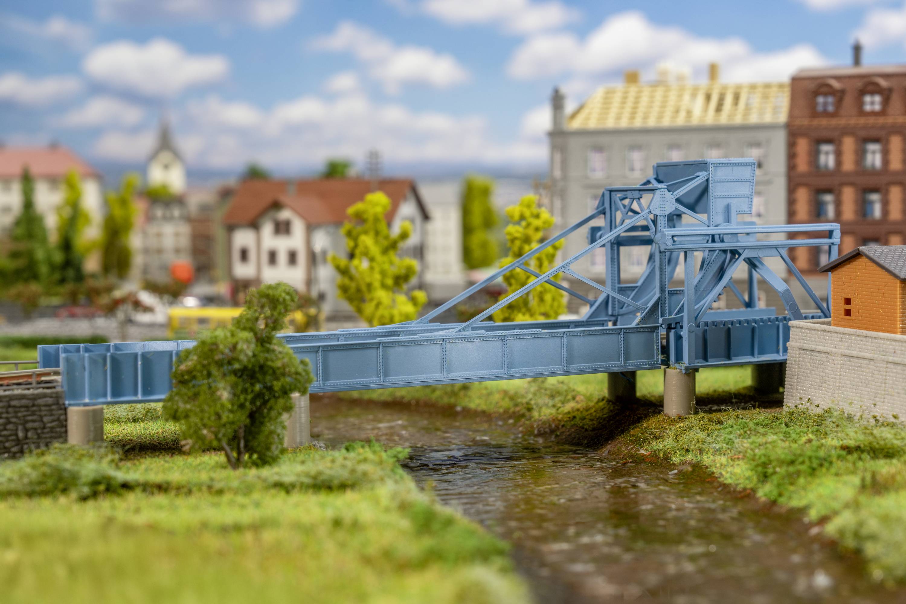 Blaues Modell einer beweglichen Brücke über einen Fluss, umgeben von Miniaturhäusern und Bäumen, mit bewölktem Himmel im Hintergrund.