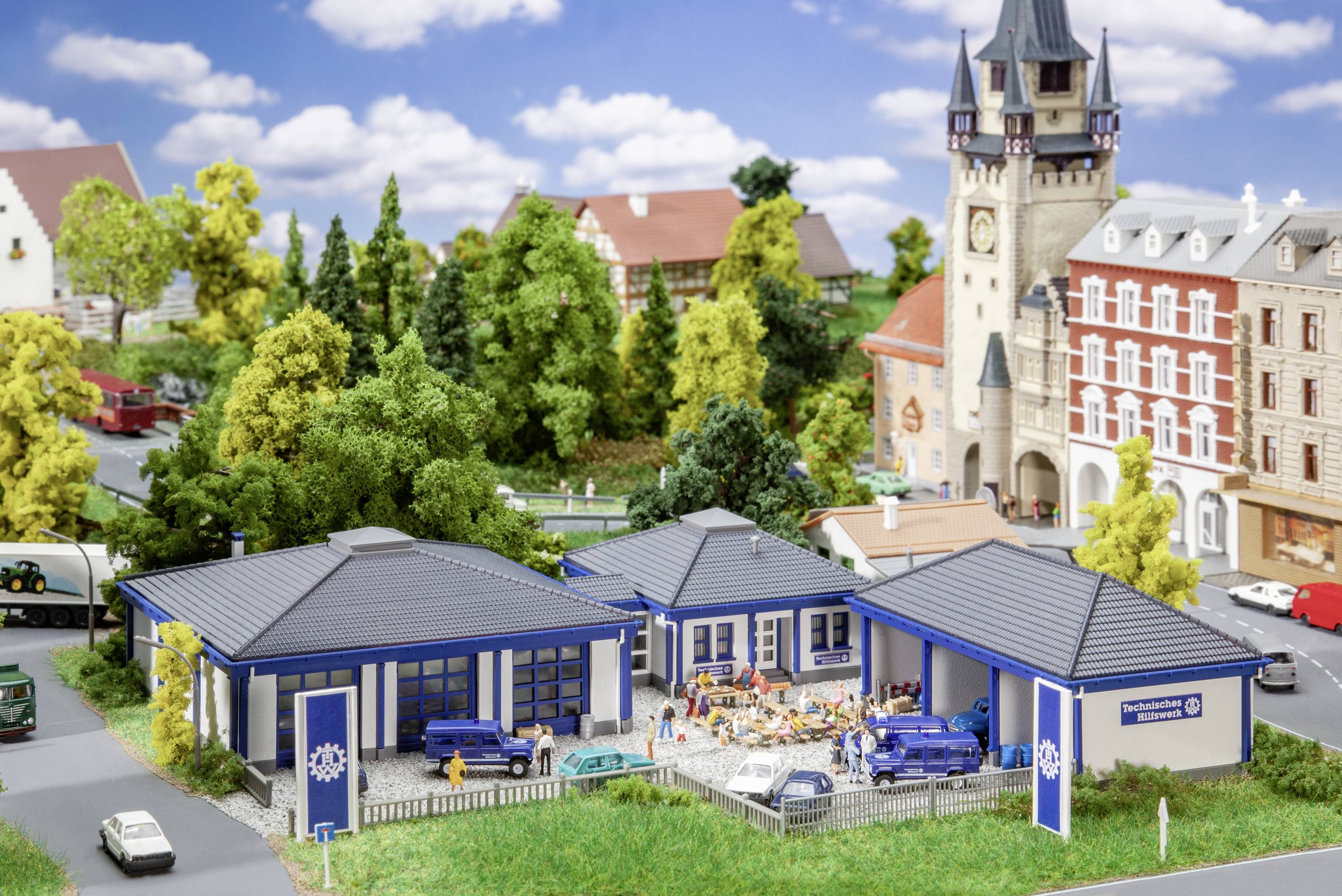 Modelleisenbahnlandschaft mit Gebäuden und Fahrzeugen, darunter ein Feuerwehrfahrzeug. Im Hintergrund ein Turm und Häuser, umgeben von Bäumen.