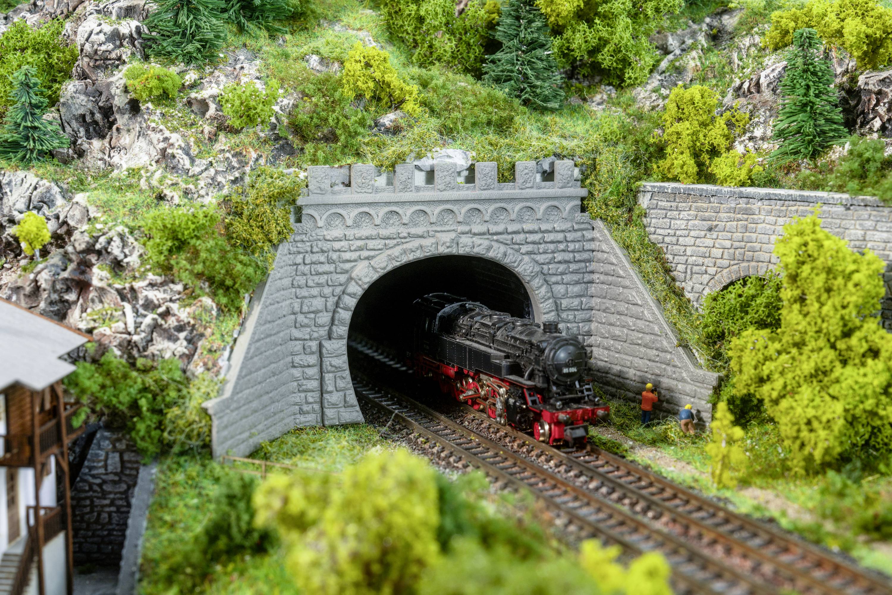 Eine Modelleisenbahn fährt aus einem Tunnel in einer bergigen Landschaft. Im Vordergrund stehen zwei Figuren neben den Gleisen.