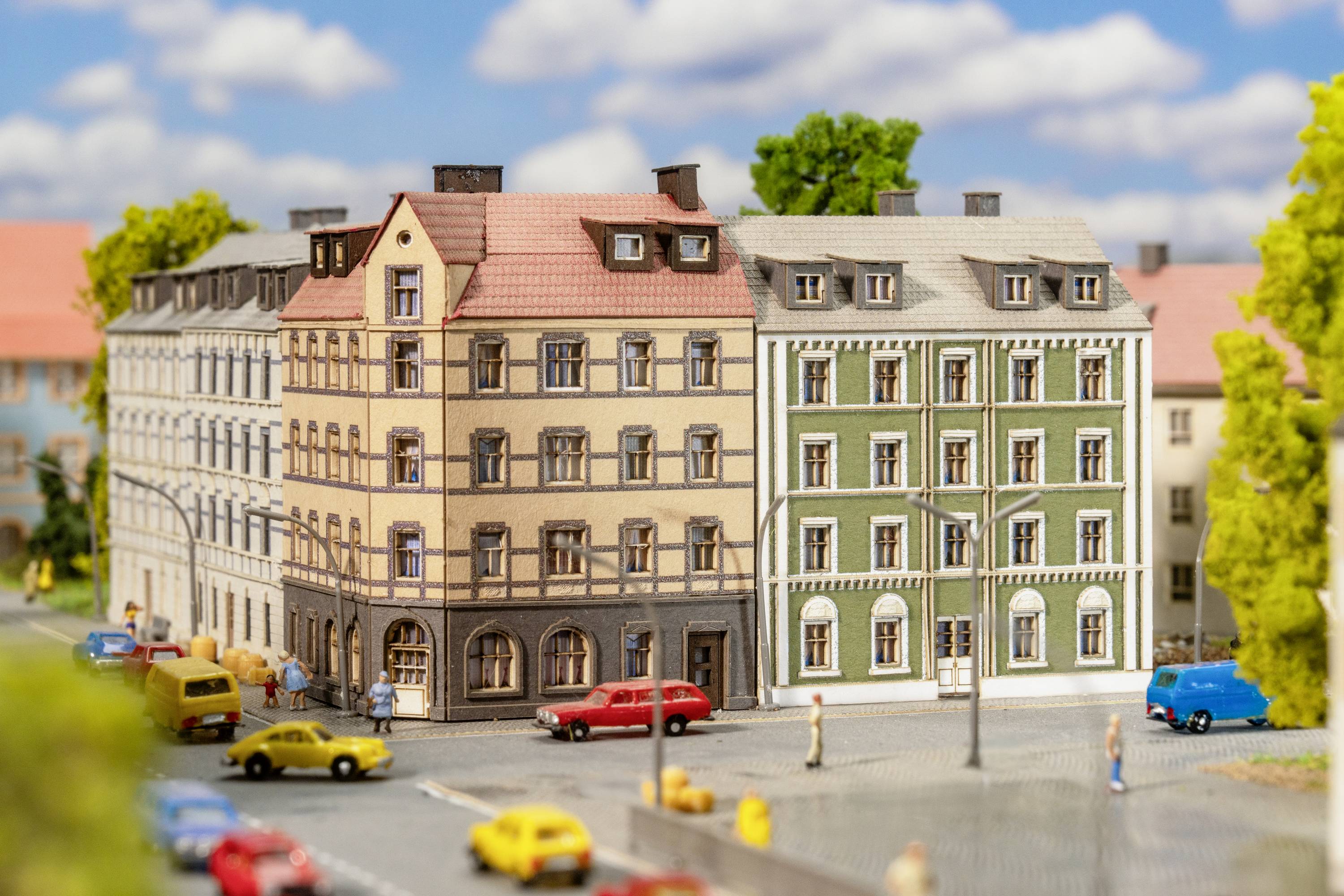 Mehrstöckige Altstadtgebäude mit roten und grünen Fassaden, Autos und Passanten auf einer belebten Straße unter blauem Himmel.