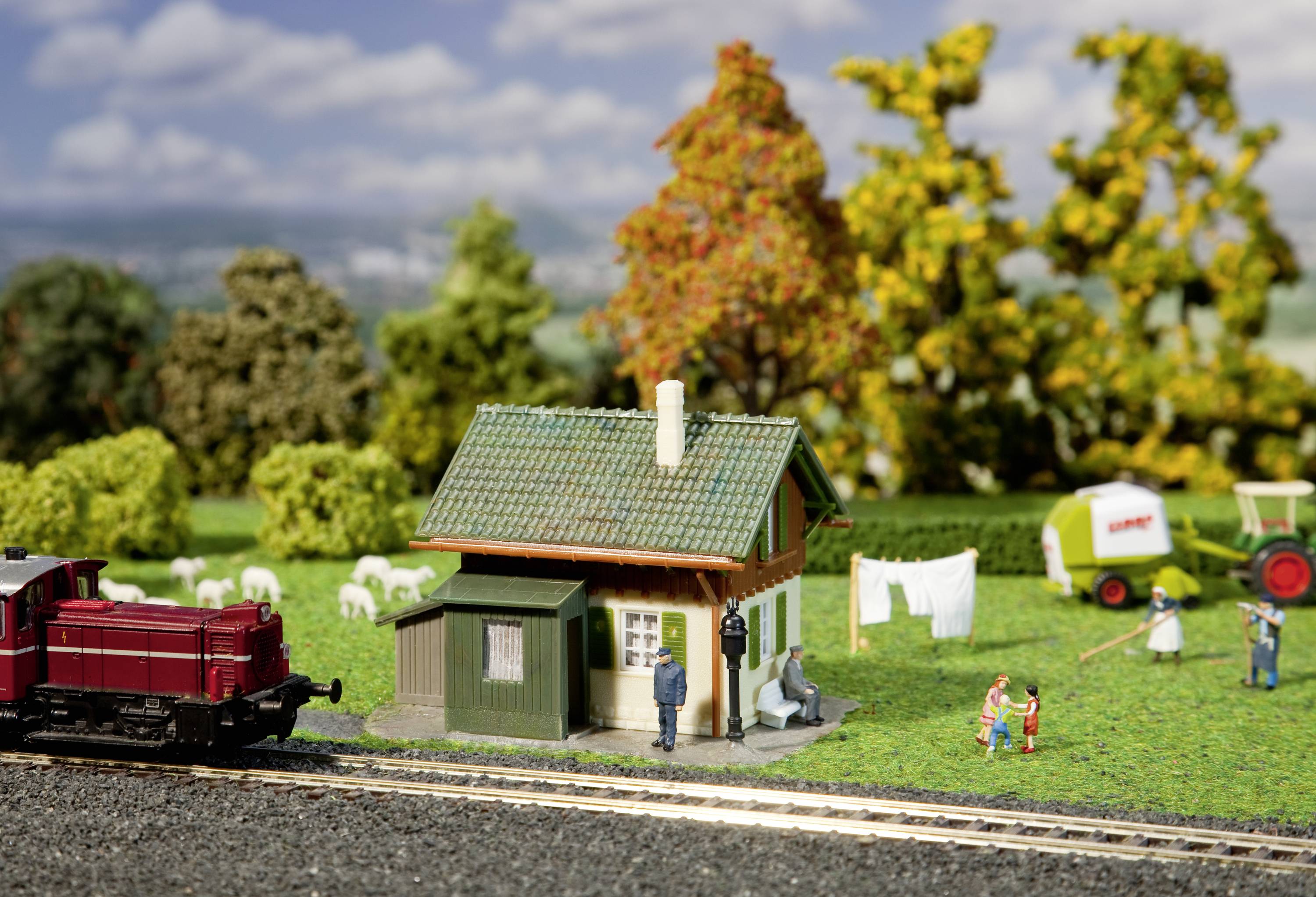 Eine Miniaturszene mit einem kleinen Haus und Wäsche an der Leine, umgeben von Bäumen. Im Vordergrund verläuft eine Modellzugstrecke.