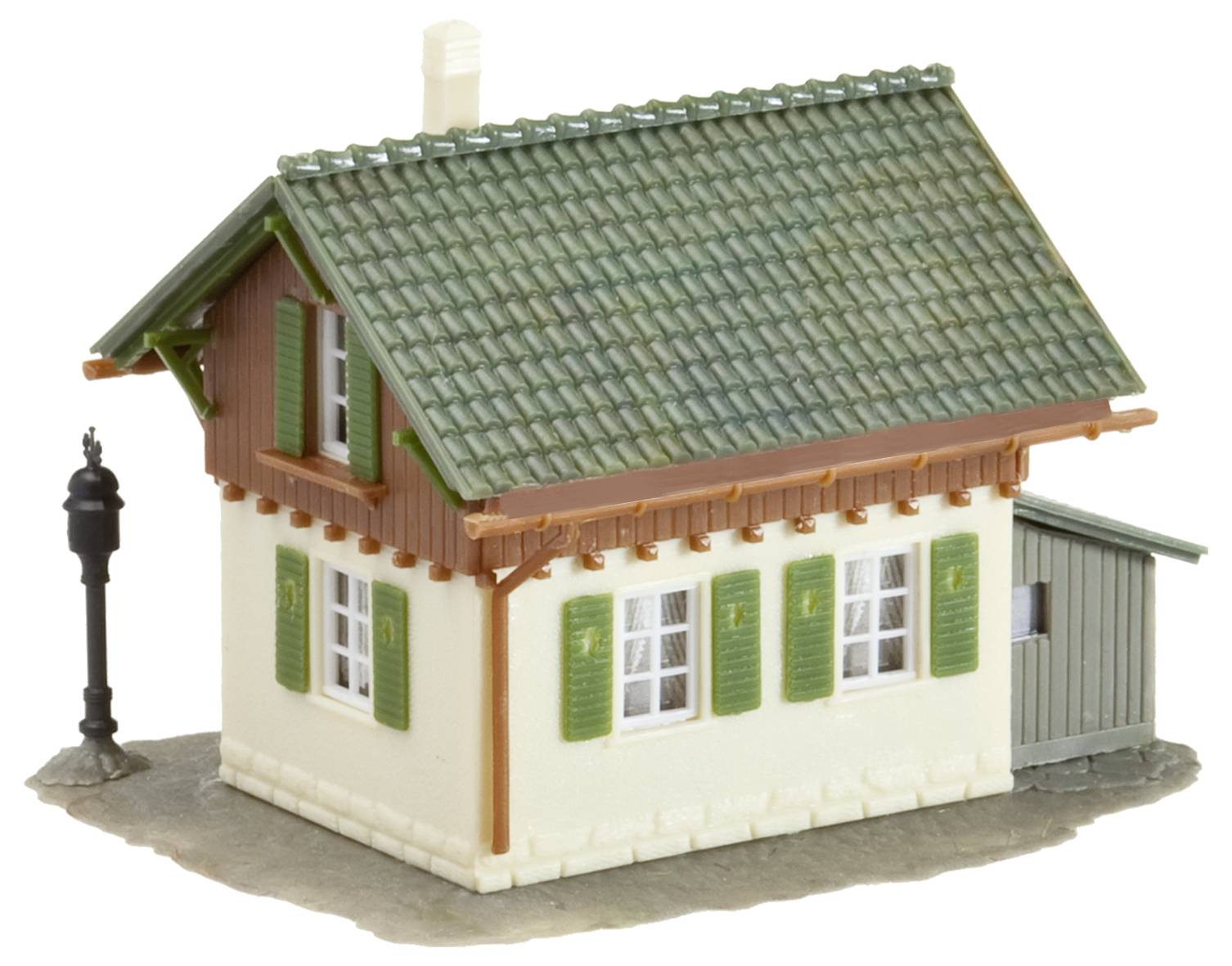Ein kleines Modellhaus mit grünem Dach, grünen Fensterläden, braunen Holzdetails und einem angrenzenden Schuppen; Laterne links.