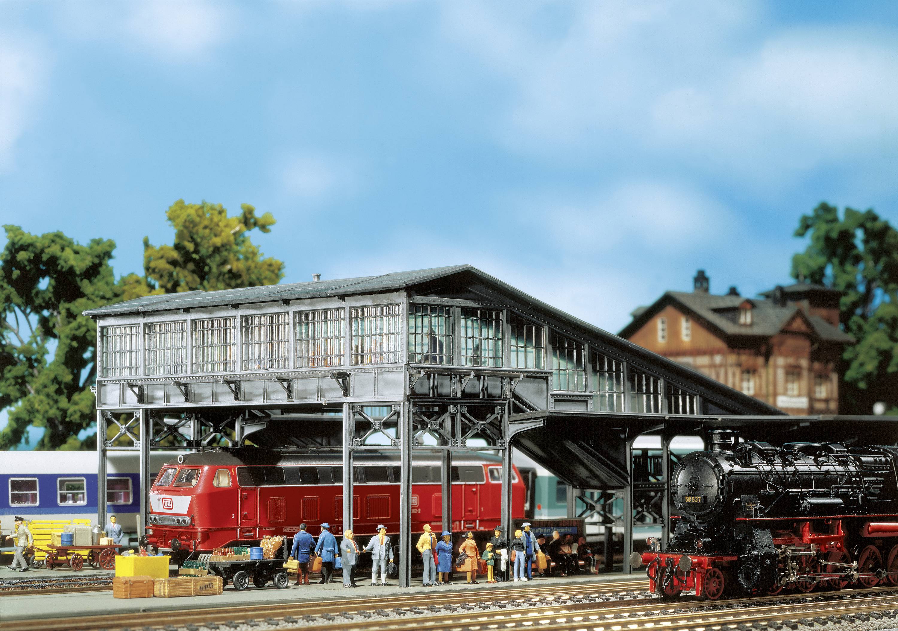 Eine Modelleisenbahnszene mit einem roten Zug unter einer Bahnwerkstatt, umgeben von Miniaturfiguren und weiteren Zügen im Hintergrund.