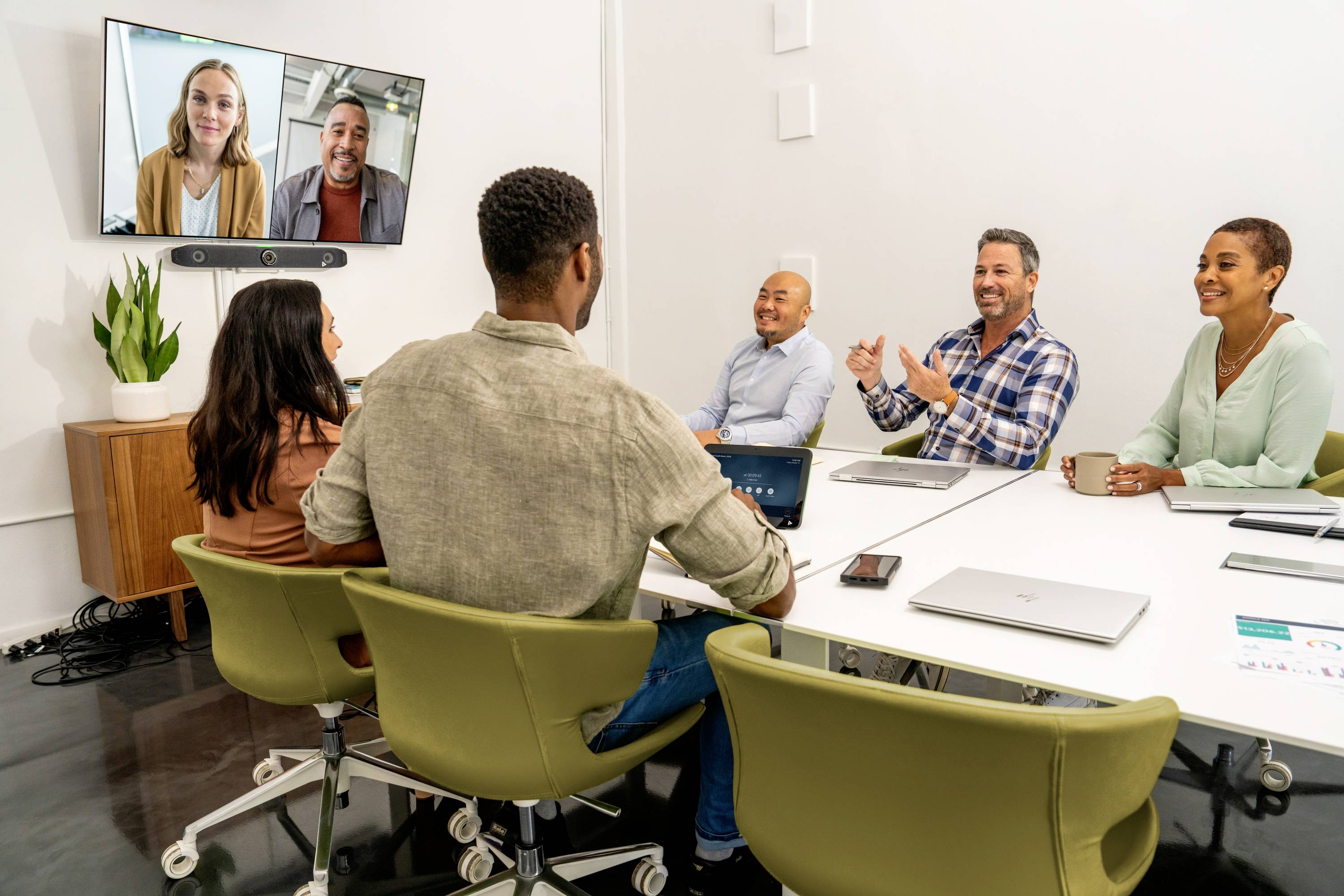 Sechs Personen in einem Meetingraum, vier am Tisch, zwei auf einem Bildschirm per Videokonferenz zugeschaltet, diskutieren lächelnd.