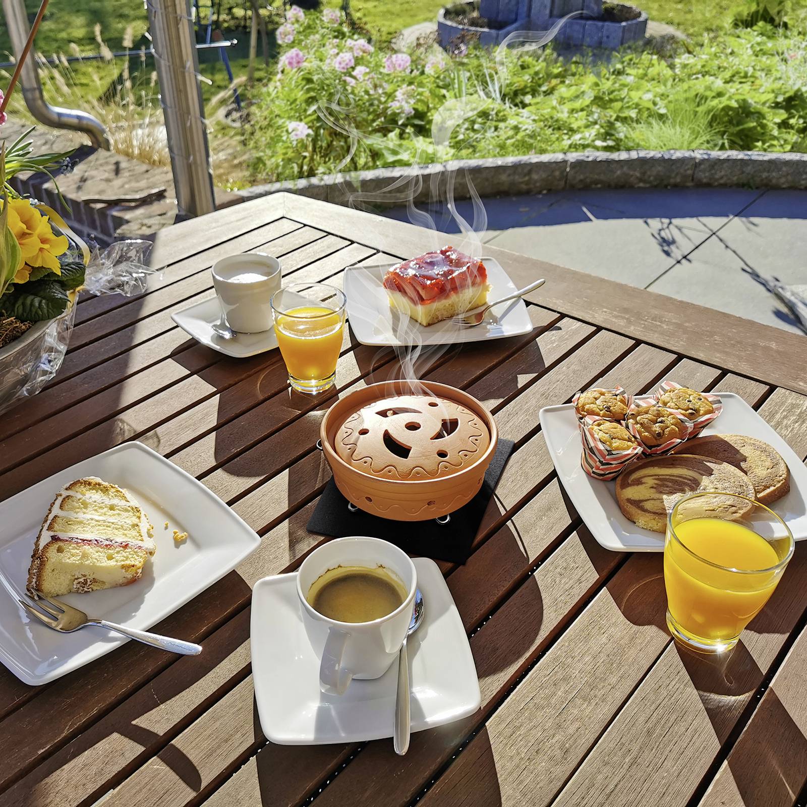 Frühstückstisch im Freien mit Kuchen, Keksen, Kaffee und O-Saft. In der Mitte rauchender Tontopf als Teelicht. Garten im Hintergrund.